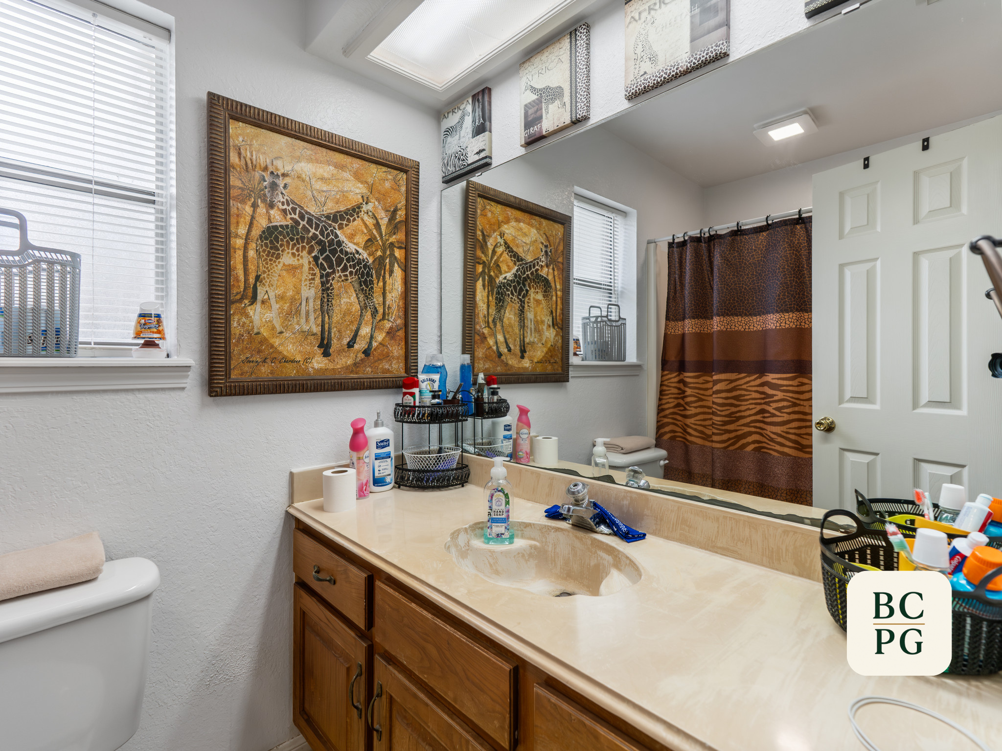 A bathroom with a large mirror, wooden vanity, and a beige countertop. On the countertop, there are various toiletries, including soap, hand sanitizer, and toothbrushes. To the left, a toilet with a beige a towel placed on top. The wall is decorated 