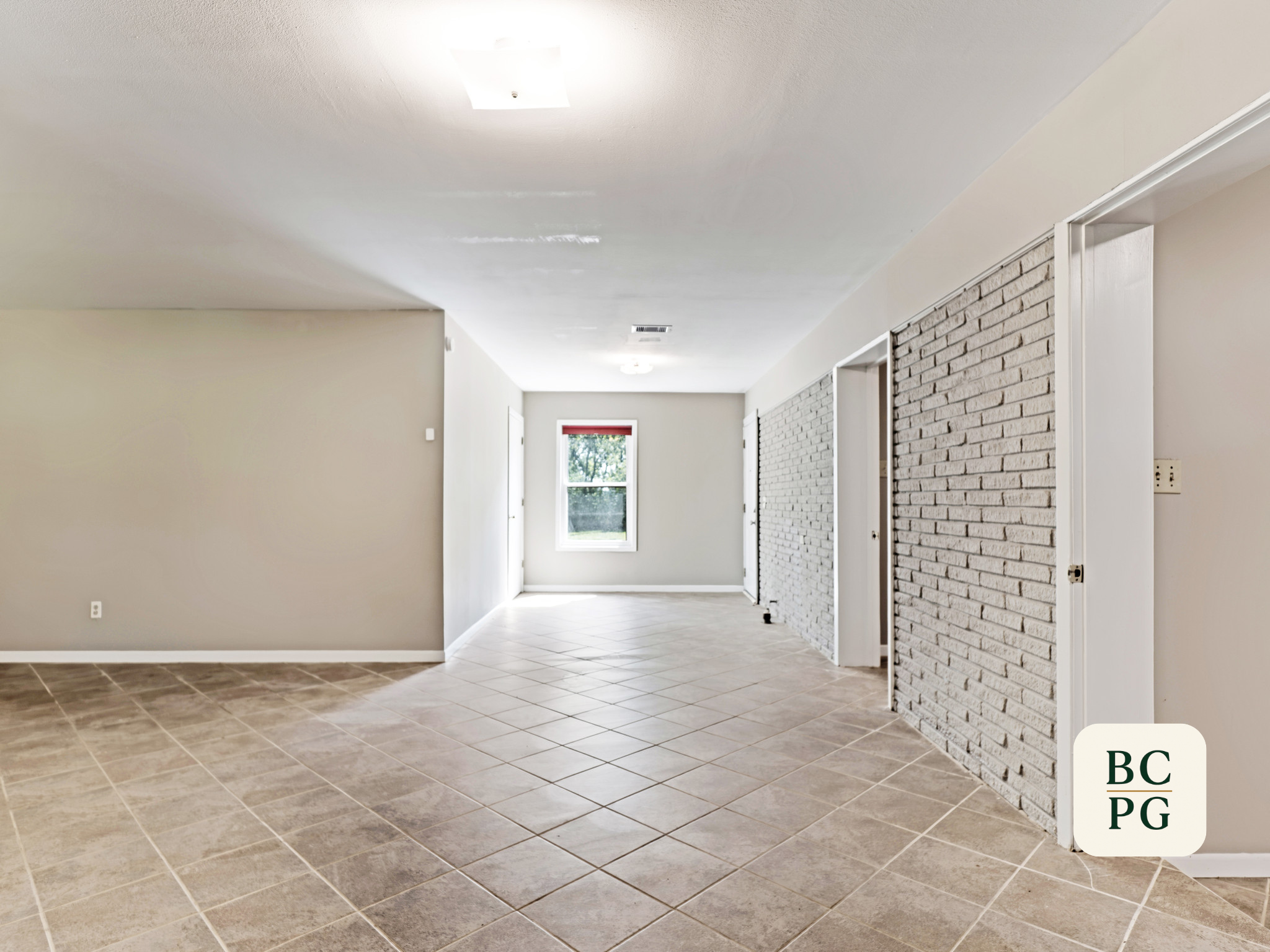 Interior view of a bright, empty hallway with beige tiles on the floor, white-painted walls, a brick accent wall on the right, a window at the end, and ceiling lights.