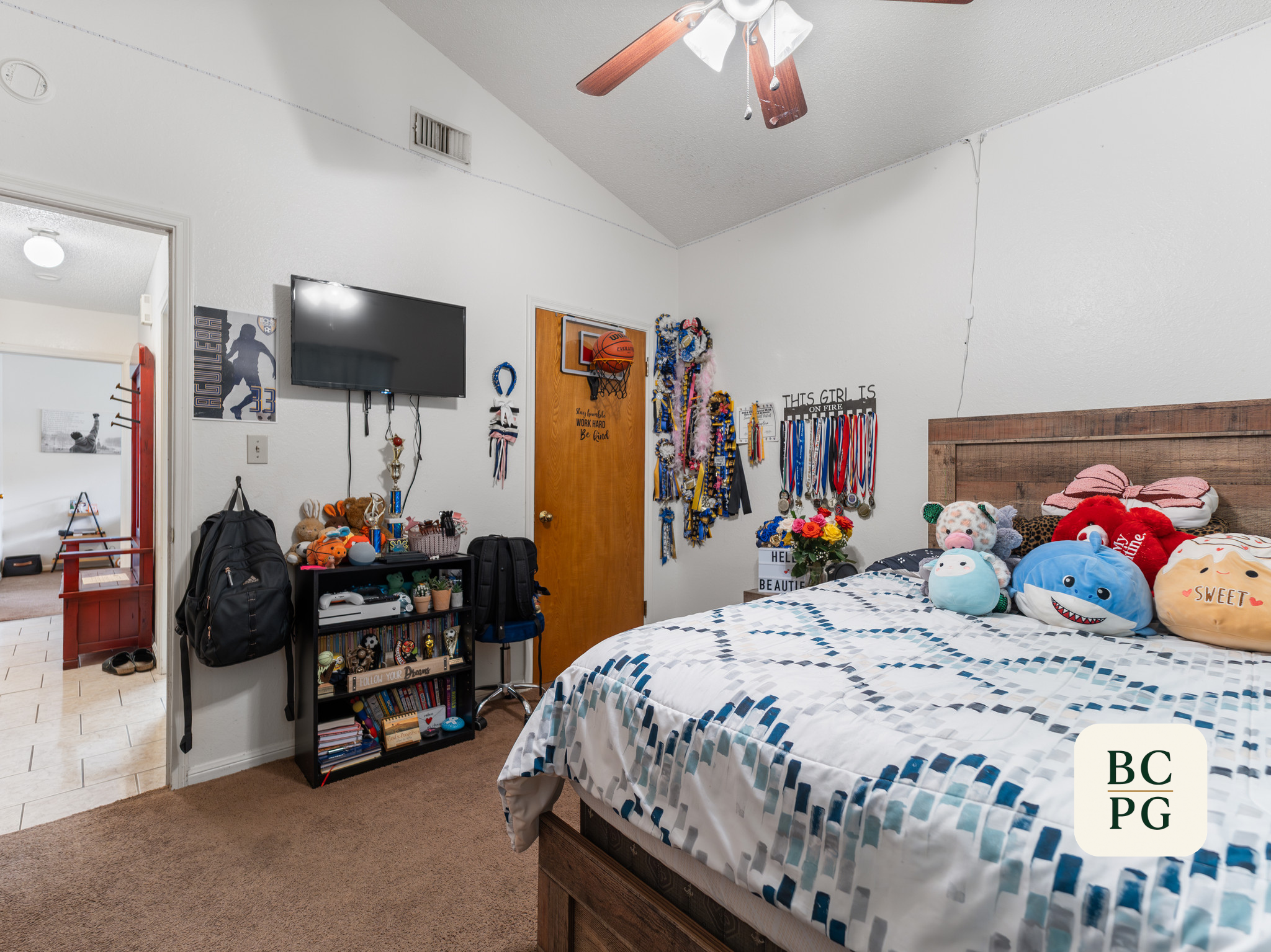 A bedroom with a bed covered in a blue, white, and gray geometric patterned quilt, stuffed toys, and pillows. The wall opposite the bed has various ribbons and medals hanging on hooks, with a sign that reads "This girl is on fire." There is a flat-sc
