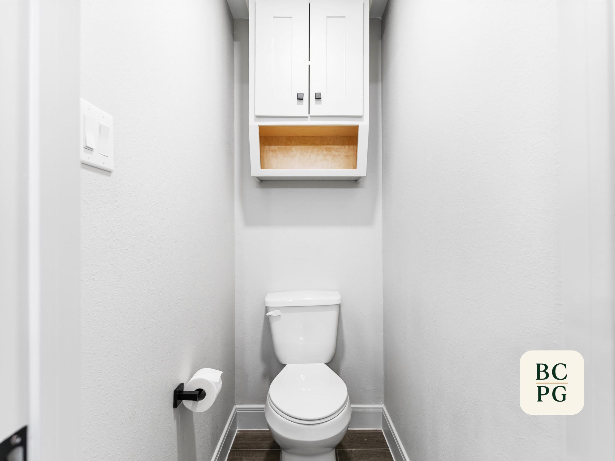 Small bathroom with white walls, a white toilet, and a wooden cabinet above the toilet. There is a toilet paper roll on a holder to the left and a light switch on the wall. The flooring is dark wood.