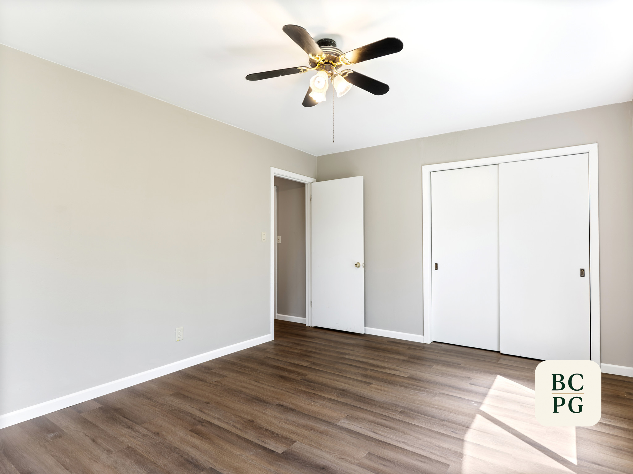 Empty room with beige walls, white ceiling, wooden flooring, ceiling fan with lights, open door, and sliding closet doors.