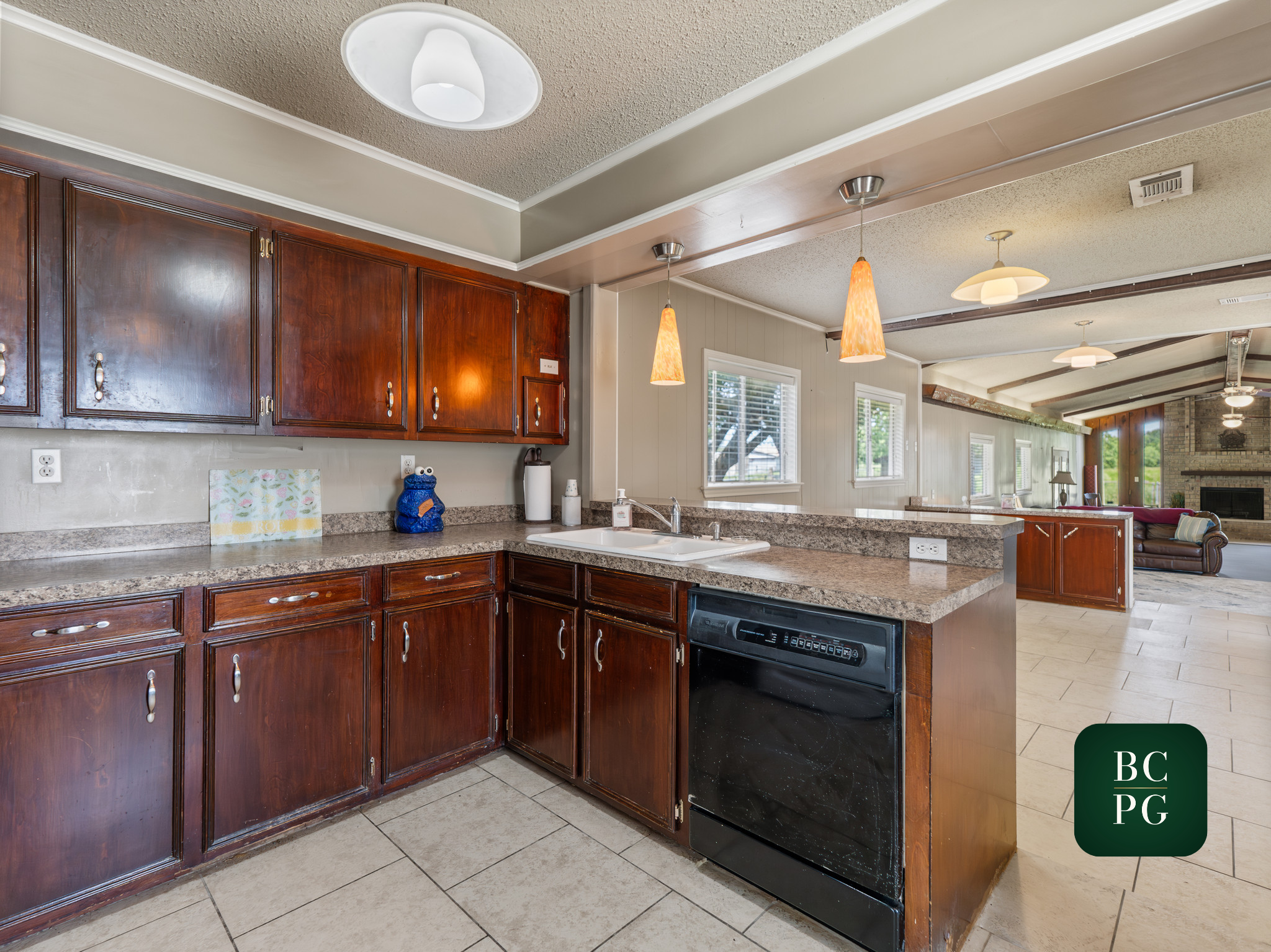 Kitchen with dark wood cabinets, granite countertop, white double sink, hanging pendant lights, and a view into a bright living room with windows and a brick fireplace.