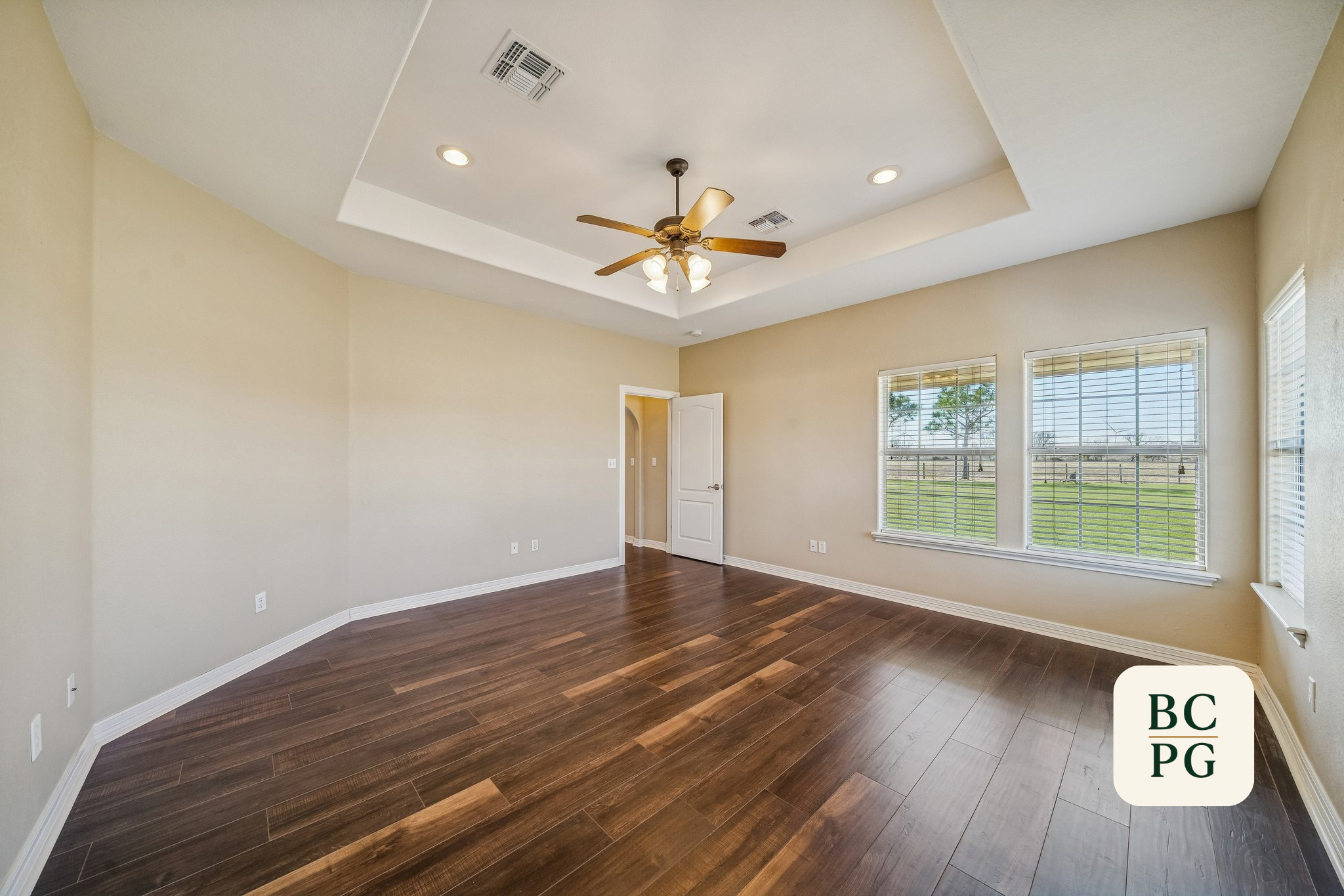Empty living room with hardwood floors, beige walls, three large windows with blinds, white door, ceiling fan with lights, and a tray ceiling design.