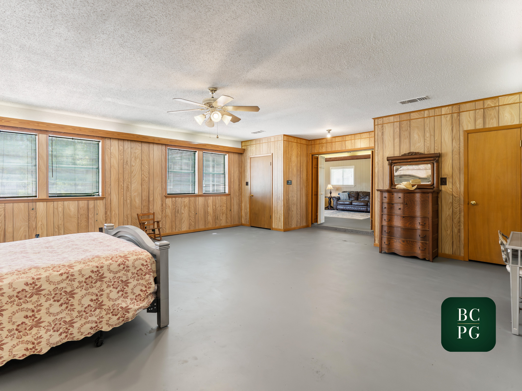 Interior view of a room with wood-paneled walls, several windows with blinds, a ceiling fan, a bed with floral bedding, and a mirror on a dresser.