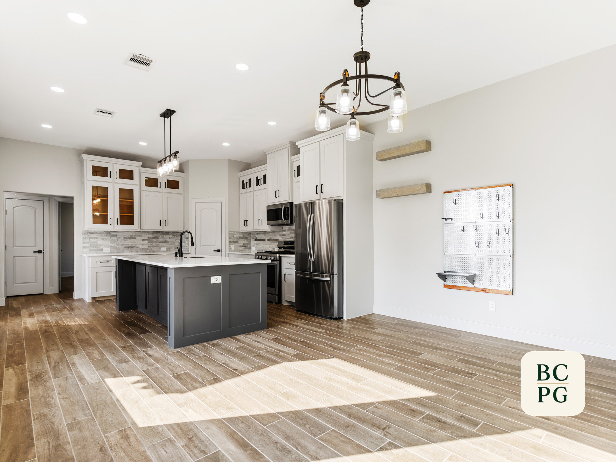 A modern kitchen with white cabinets, a kitchen island with a dark gray base, stainless steel appliances, and hanging light fixtures. The floor has wood-look tile, and there are two floating shelves on the wall. The ceiling is white with recessed lig