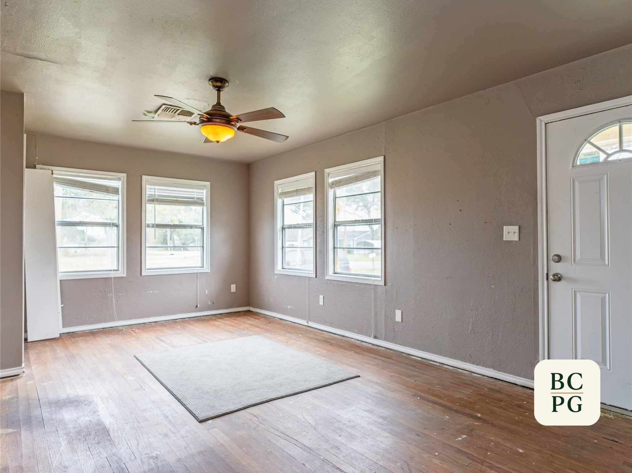 Empty living room with hardwood floors, three large windows, gray walls, a ceiling fan with light, and a white front door.