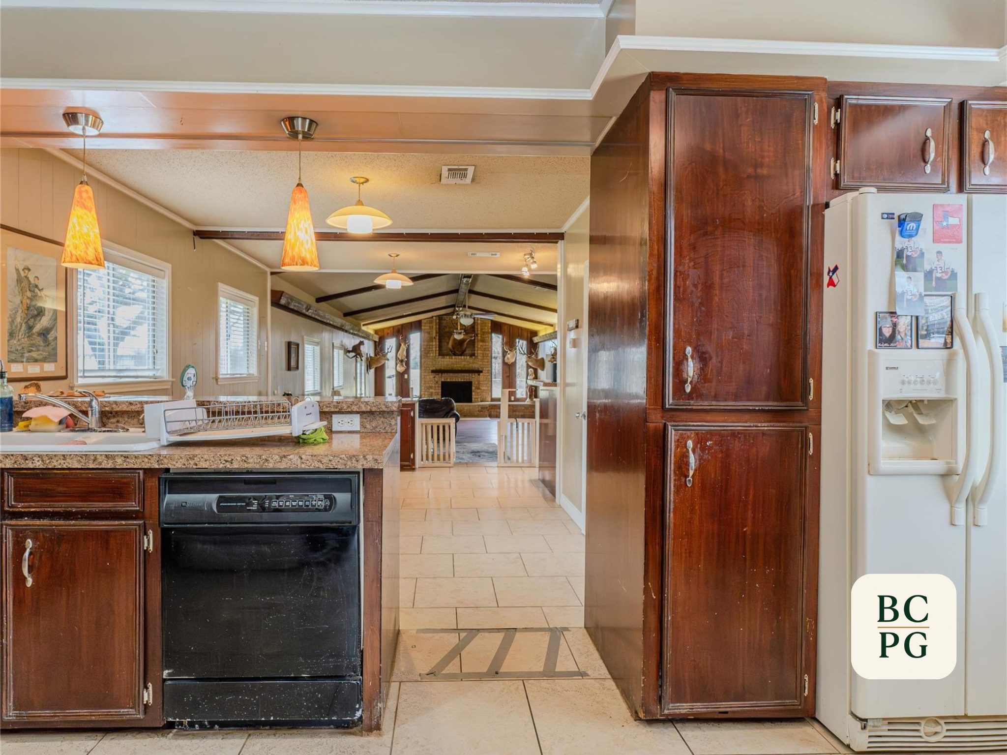 Interior view of a kitchen with wooden cabinets, a white refrigerator, tiled floor, and decorative mounted animal heads in the background.