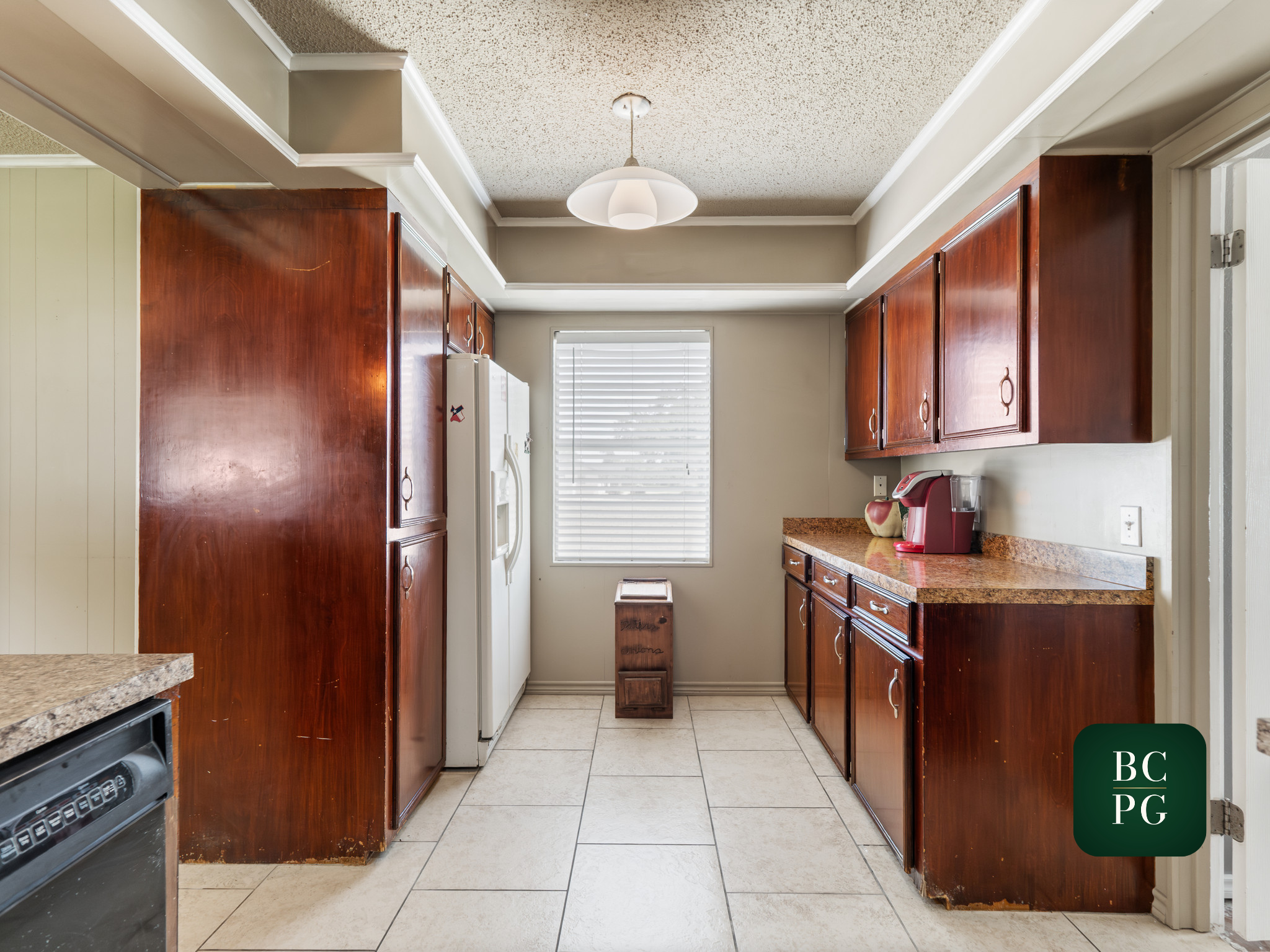 View of a kitchen with wooden cabinets, a white refrigerator, a window with blinds, beige tiled floor, and a ceiling light fixture.