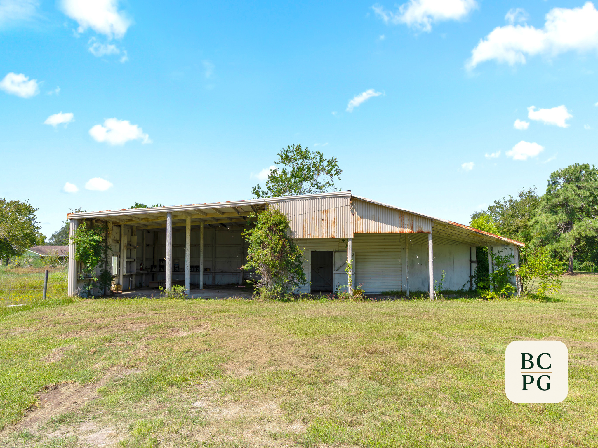 A large open metal building with a rusted roof, surrounded by green trees and grass, under a bright blue sky with scattered white clouds.