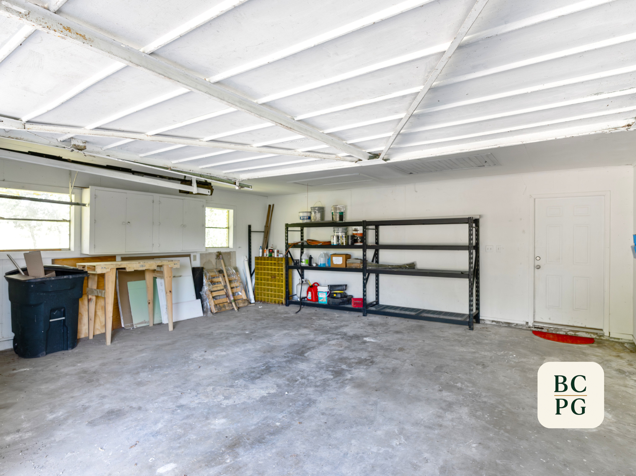 Empty garage with unfinished concrete floor, metal shelving with supplies, and a white wall with windows