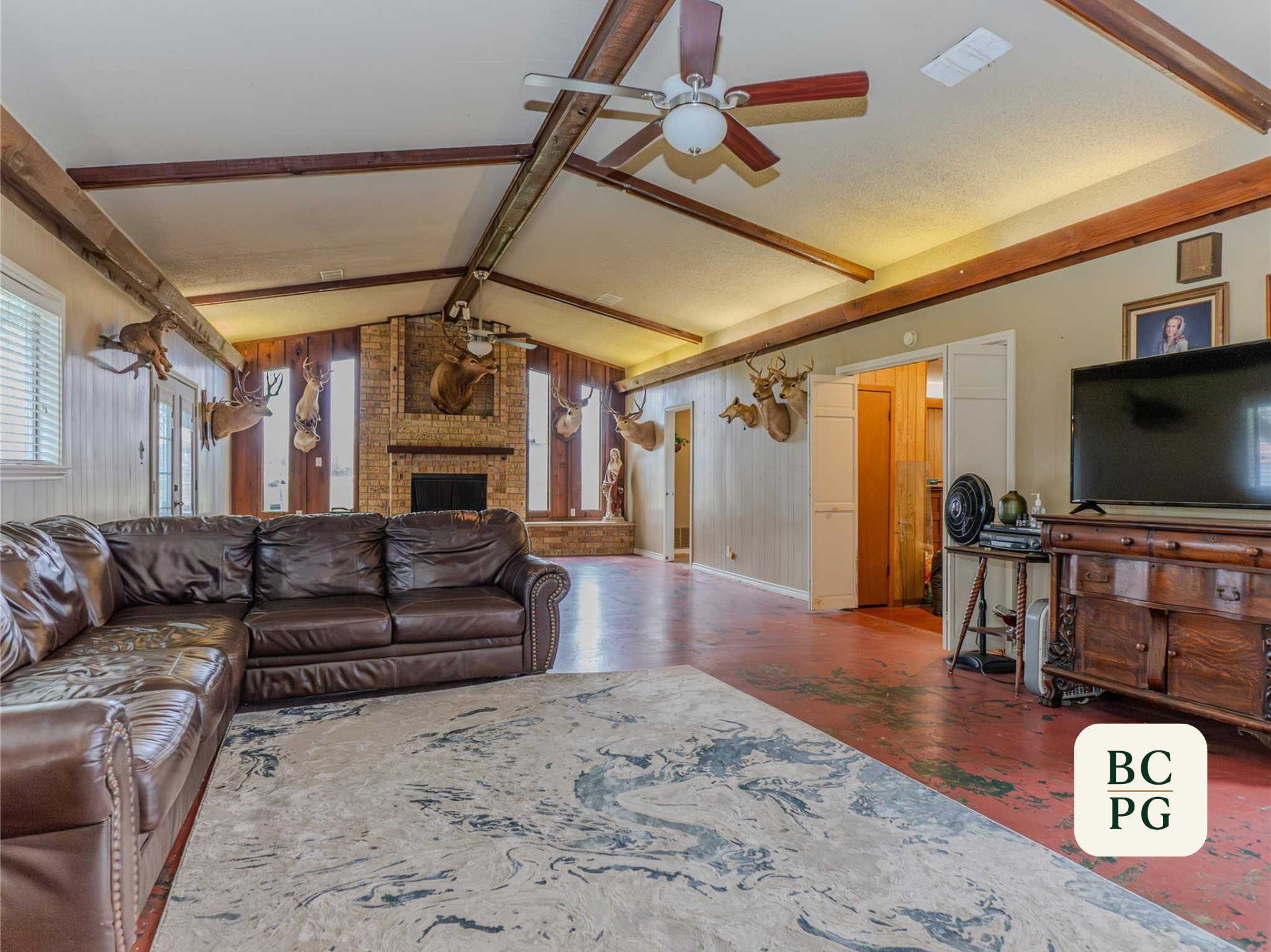 Living room with a brown leather sofa, a white area rug, and a fireplace with an animal mount above it. Walls are paneled and decorated with mounted animal heads, and there is a ceiling fan with wooden blades.