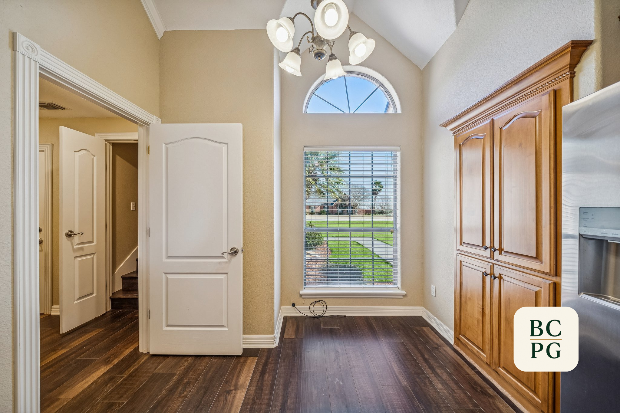 Interior view of a kitchen with a large window showing a grassy yard and palm trees outside, a wooden cabinet on the right, and a white door open to stairs on the left, with a ceiling light fixture.