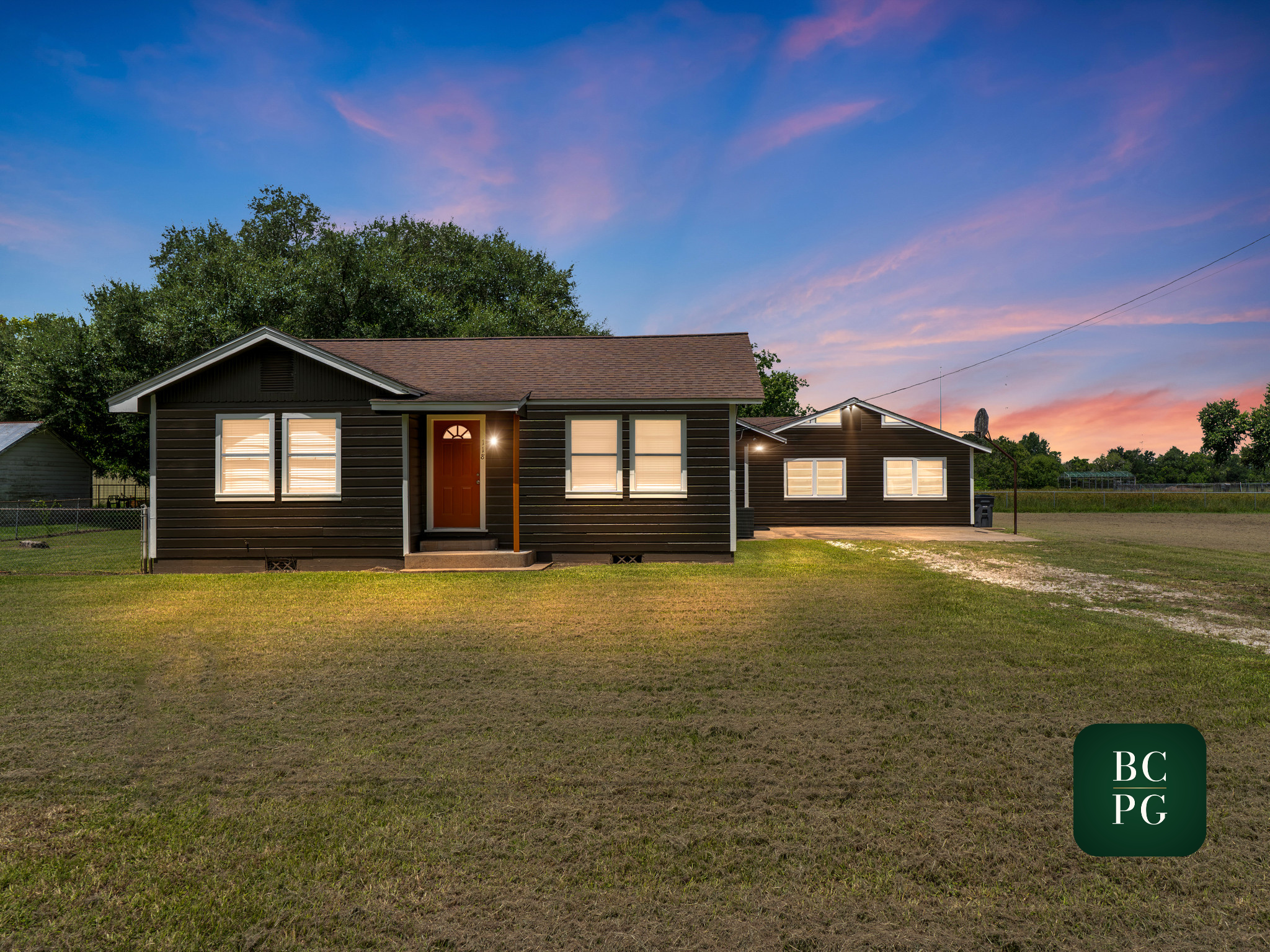 A dark brown house with a red front door and four windows, surrounded by a grassy lawn and large trees in the background during sunset.