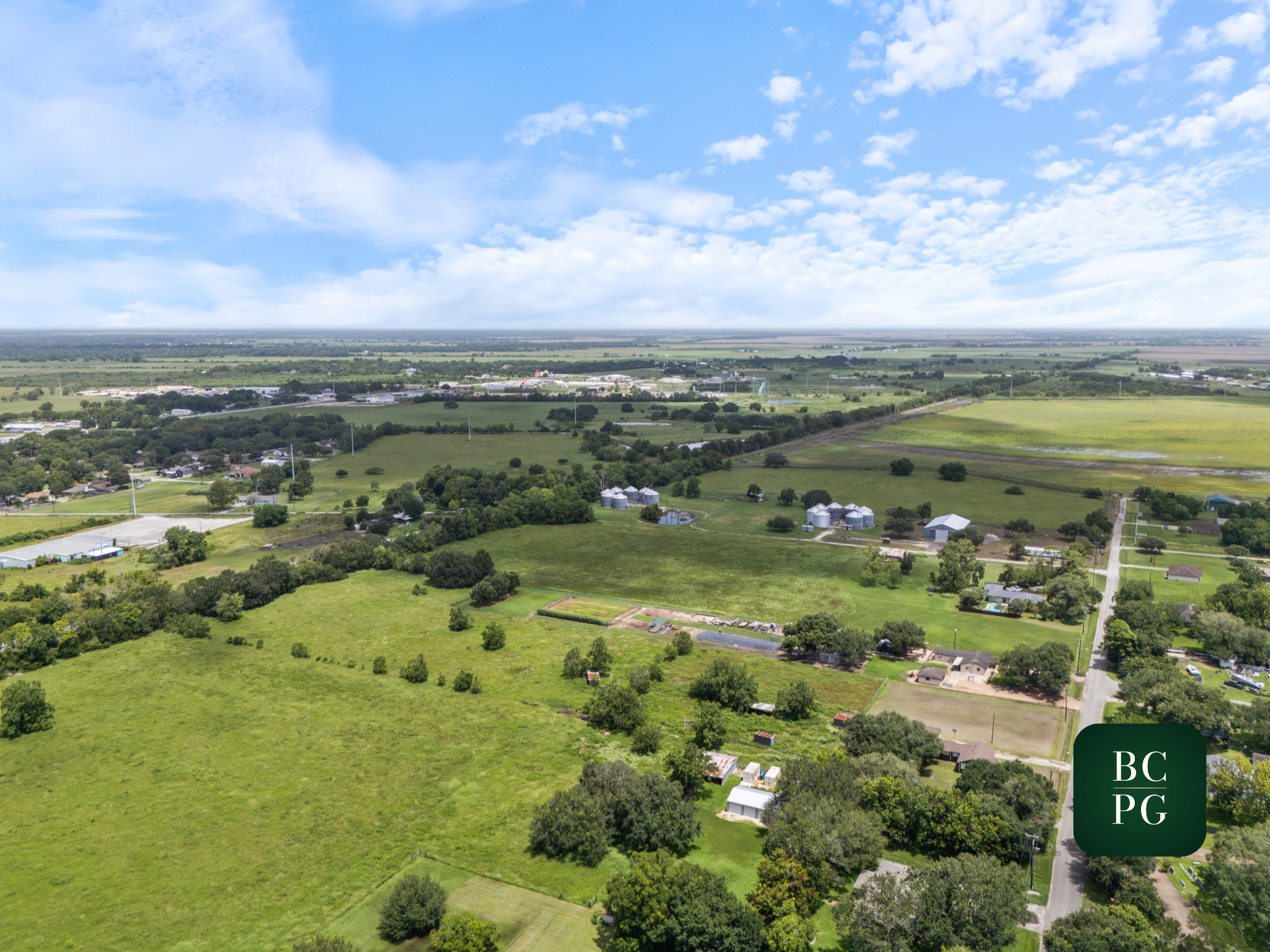 Aerial view of a rural landscape with green fields, scattered houses, trees, and a distant horizon under a partly cloudy sky.