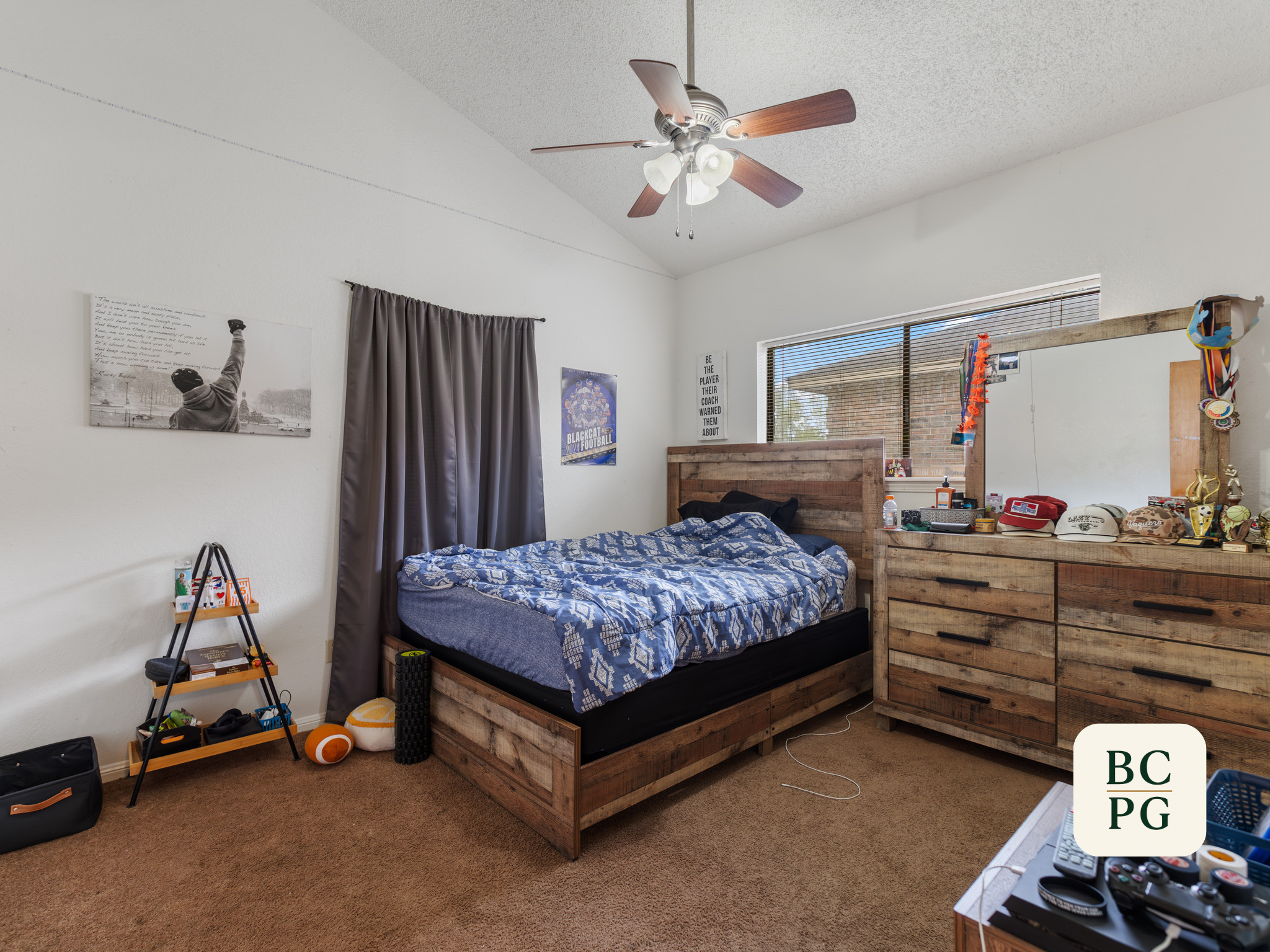 A bedroom with a ceiling fan and light fixture, a bed with patterned blue bedding and wooden frame, a dresser with medals and trophies, posters on the wall, and a window with blinds.