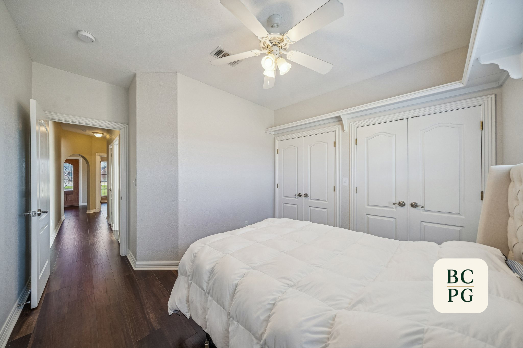 Interior of a bedroom with white walls, white closet doors, a ceiling fan with lights, a bed with white bedding, and an open door leading to a hallway with dark wood flooring and a yellow wall.