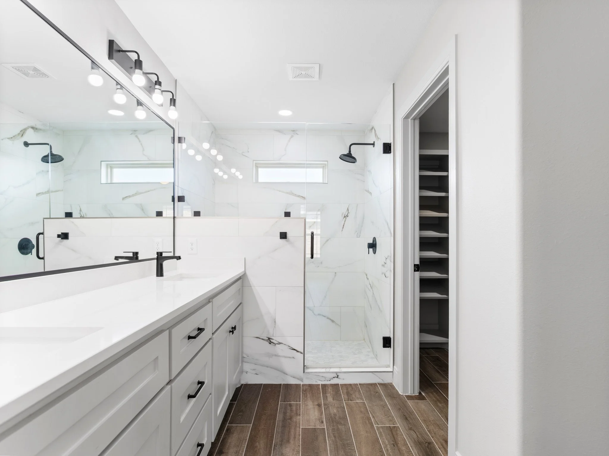 Modern bathroom with white vanity, black fixtures, large mirror, walk-in shower with marble tiles, wooden flooring, and a linen closet.