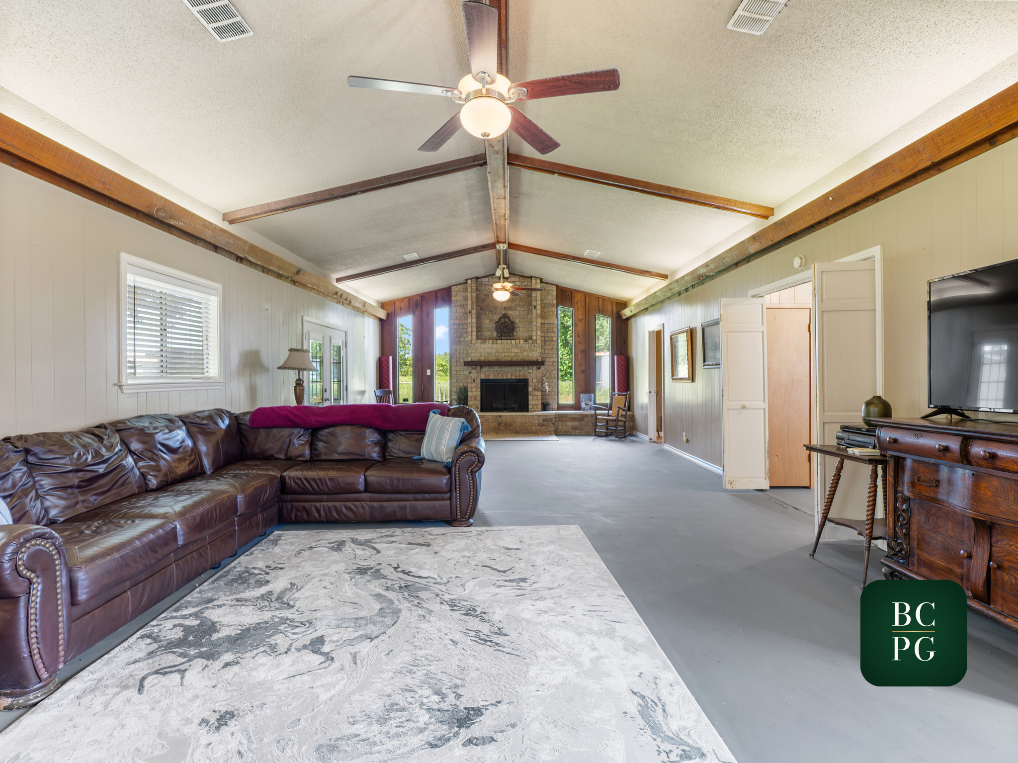 Living room with a brown leather couch, a coffee table, a fireplace, and a ceiling fan with five blades. Large windows and a sliding glass door provide natural light. The walls are paneled, and a patterned area rug is on the floor.