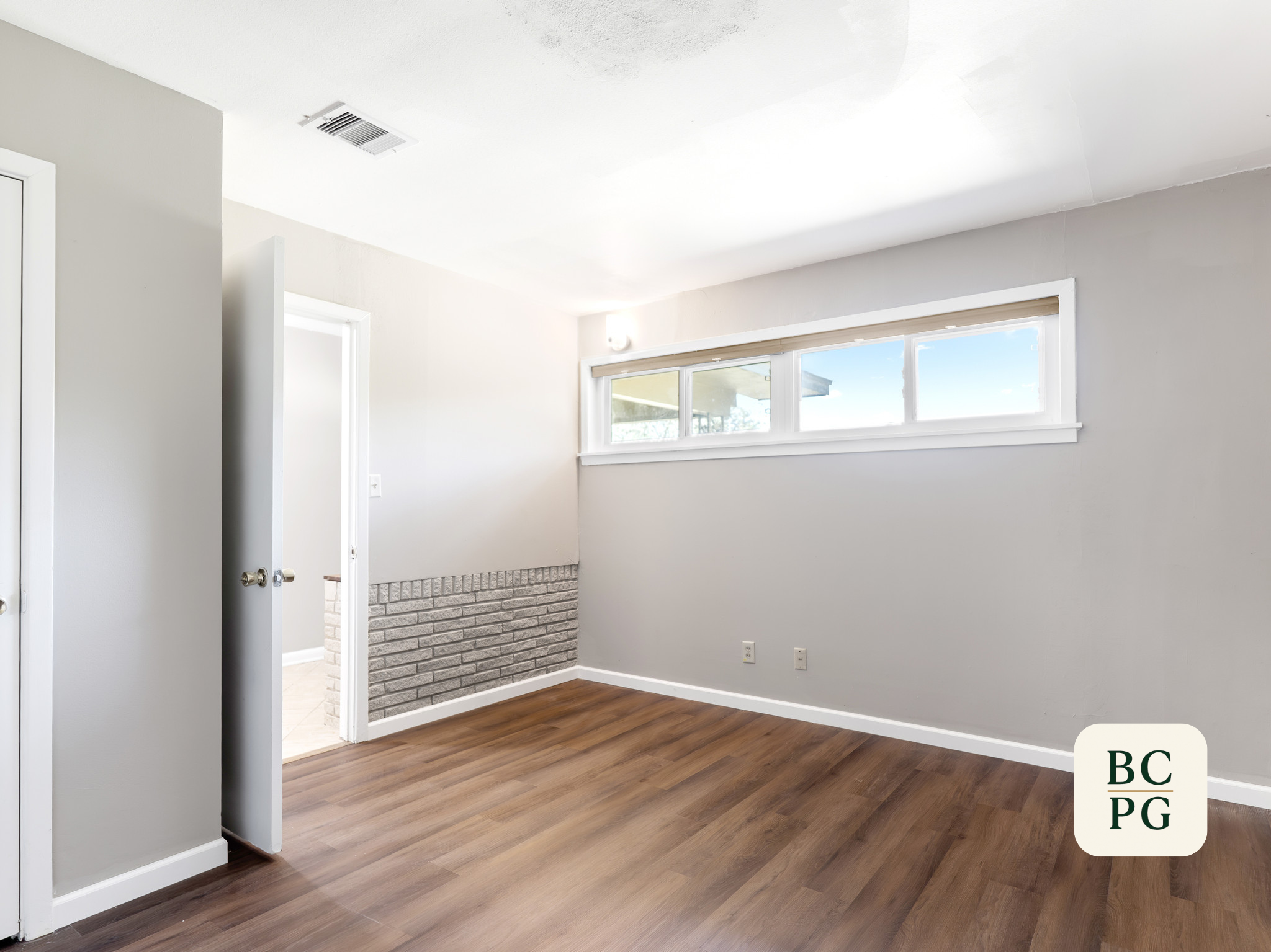 Interior of a room with white walls, wood flooring, a window with a blind, and a doorway leading to an outside area with brick wall and tiled floor.