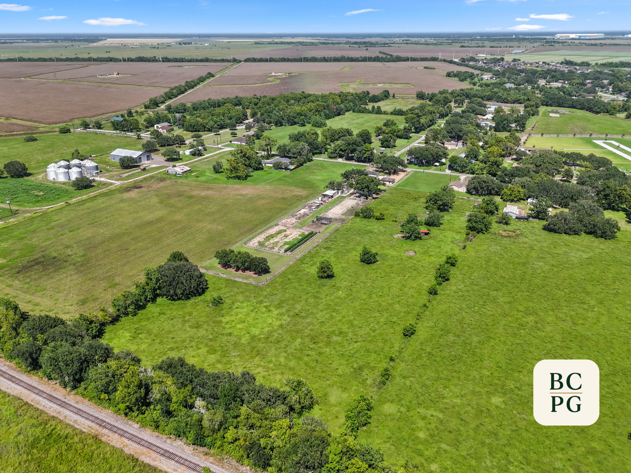 Aerial view of rural farmland with houses, trees, open fields, and a railroad track at the bottom. Distant horizon with blue sky and scattered clouds.