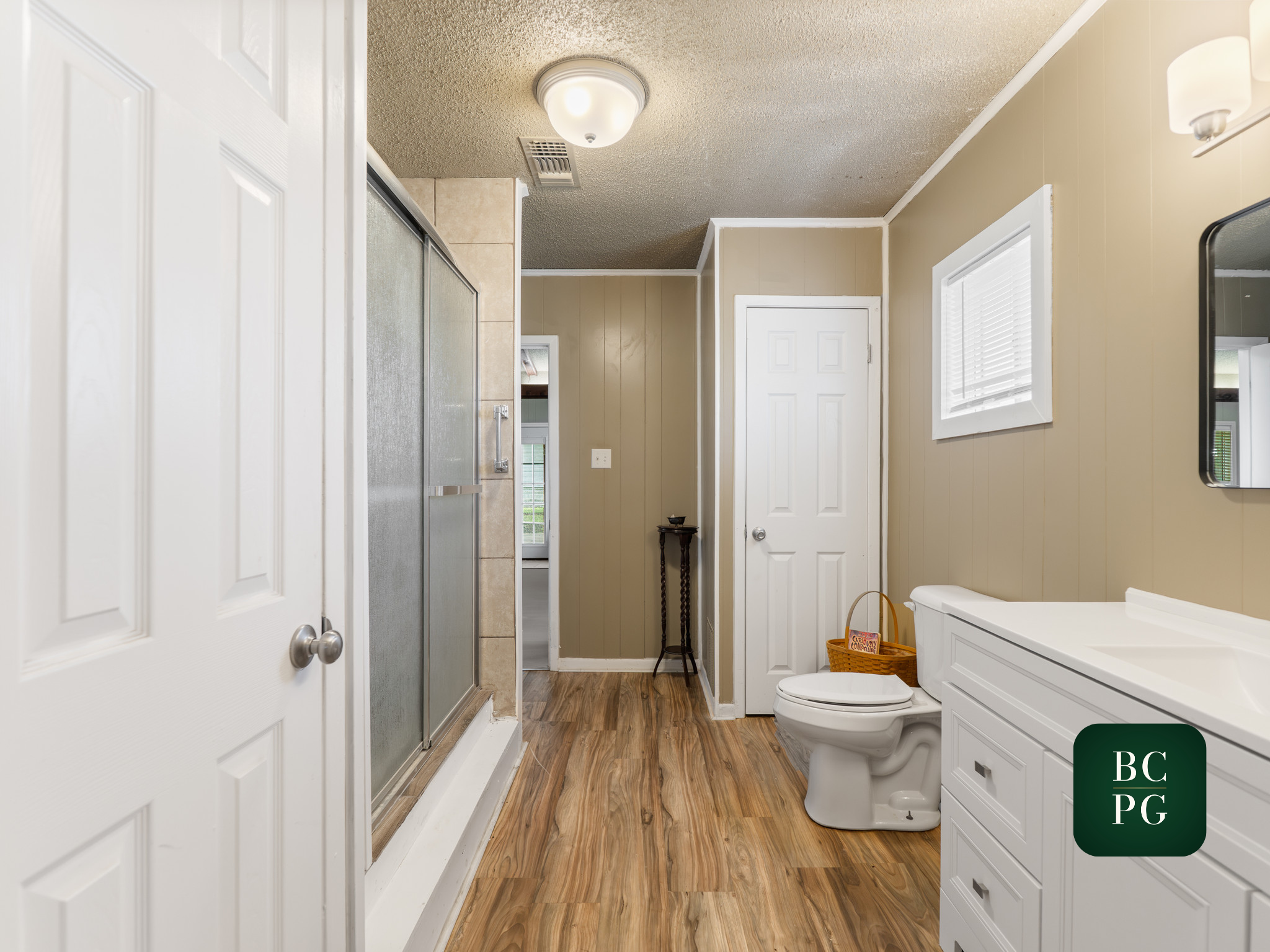 A bathroom with a white toilet, a white vanity with drawers, a wicker basket on the floor, a small black side table, and a window with blinds. There is a glass shower with a sliding door, beige tiles, and a ceiling light fixture. The walls are beige,