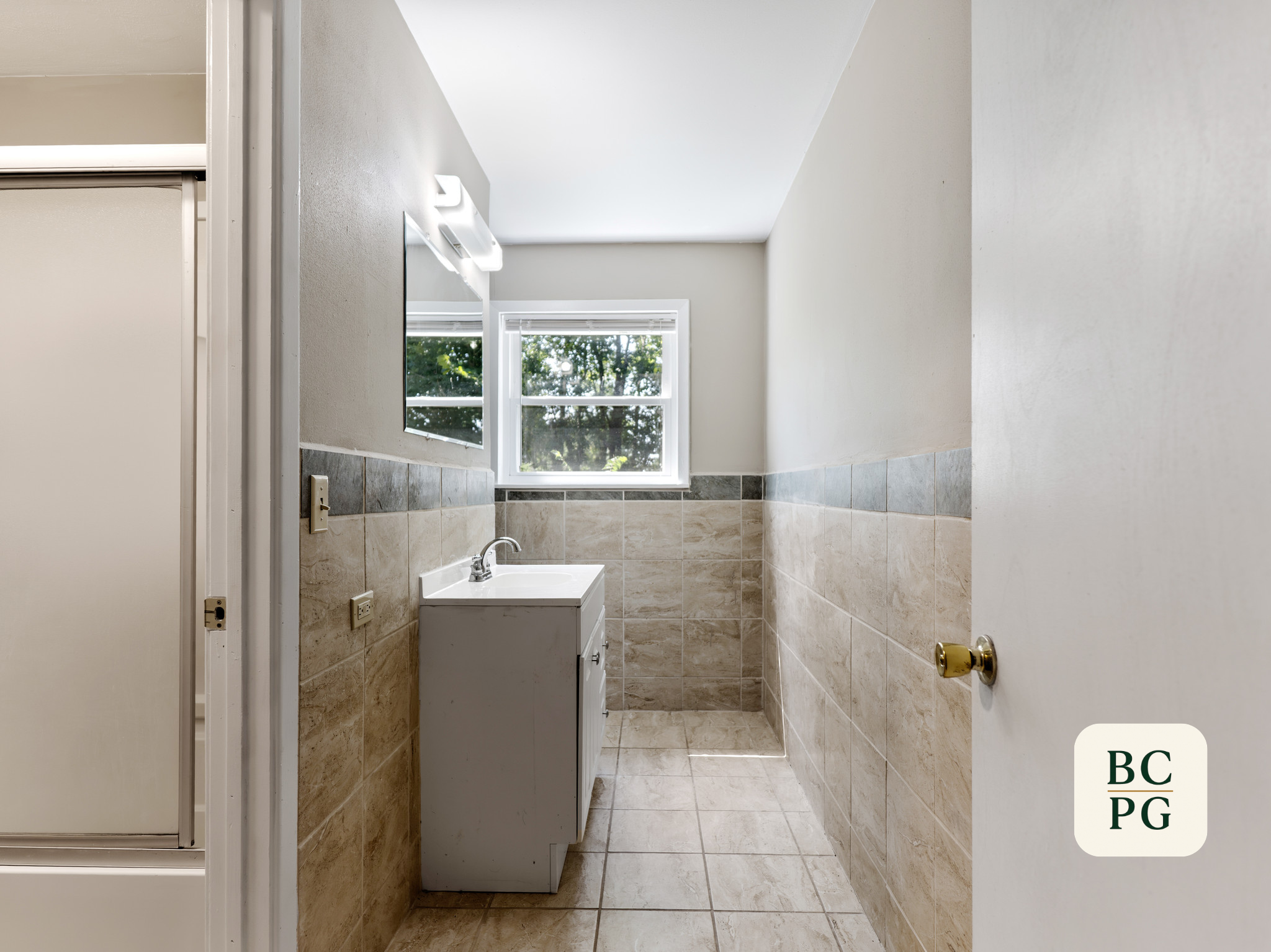 A small bathroom with beige tiled walls and floor, a white vanity with a sink, a mirror above it, and a window showing green trees outside.