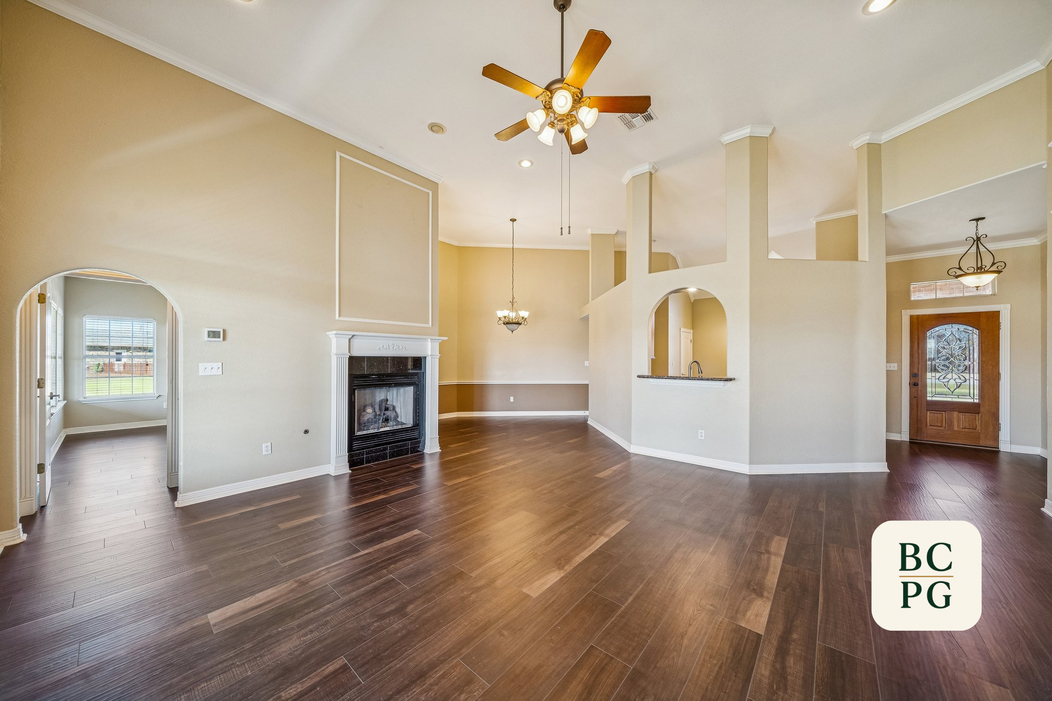Interior of a spacious living room with beige walls, white crown molding, a ceiling fan, hardwood floors, a fireplace, and multiple lighting fixtures.