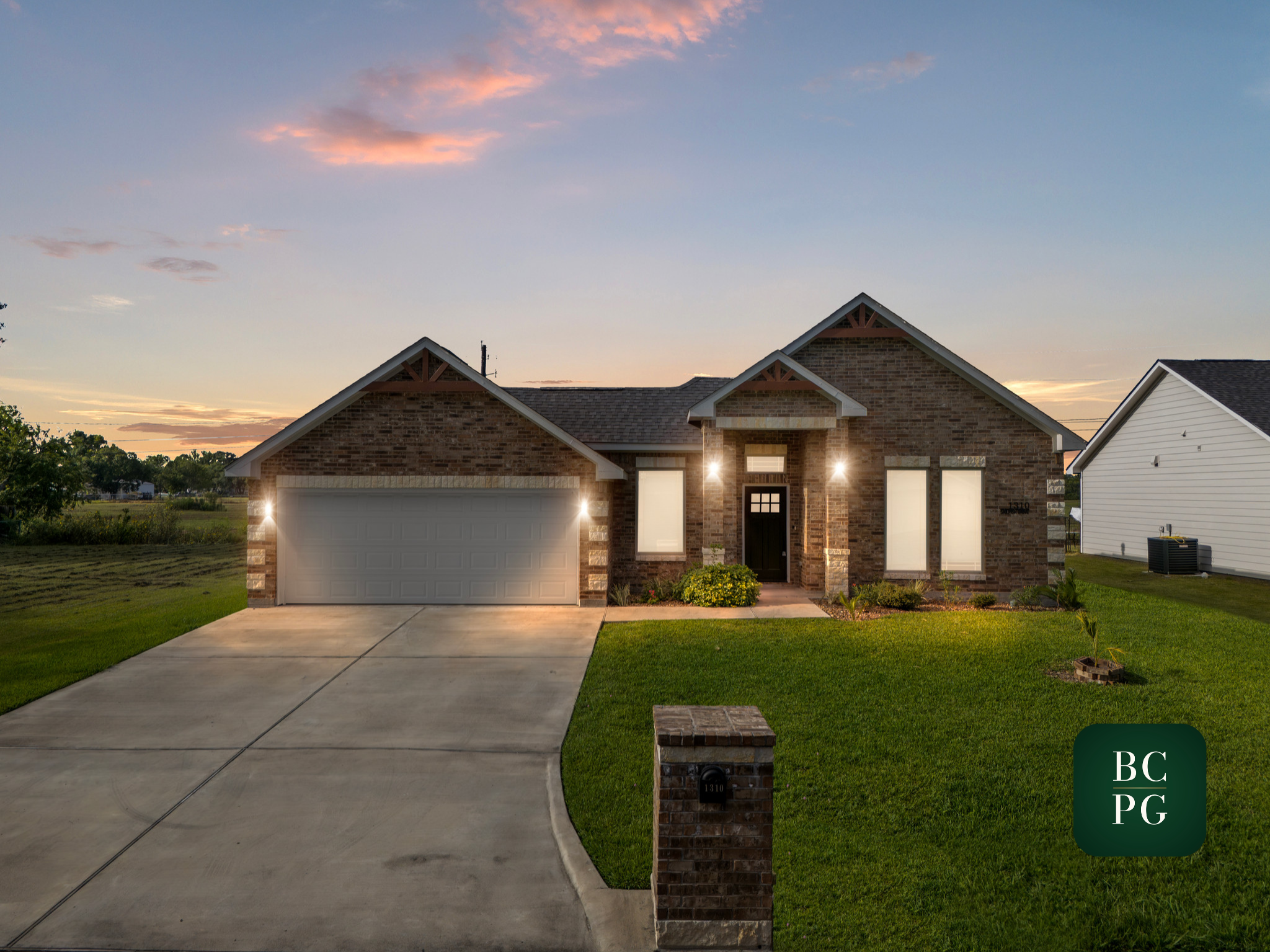 Front view of a modern brick house at sunset, with a concrete driveway, green lawn, and a small brick mailbox with house number 1310. The sky has some pink and orange clouds.