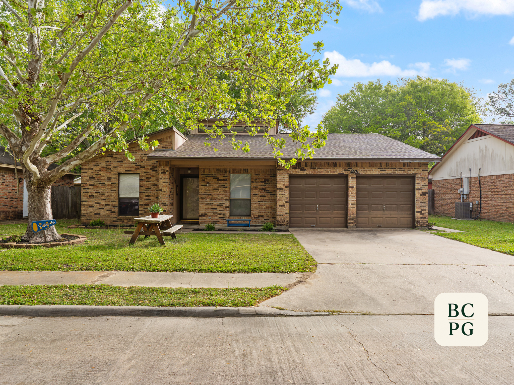 Front view of a brick house with a garage, front door, and windows. A large tree with lush green leaves is in the front yard. There is a wooden bench and potted plant near the house. The sky is partly cloudy.