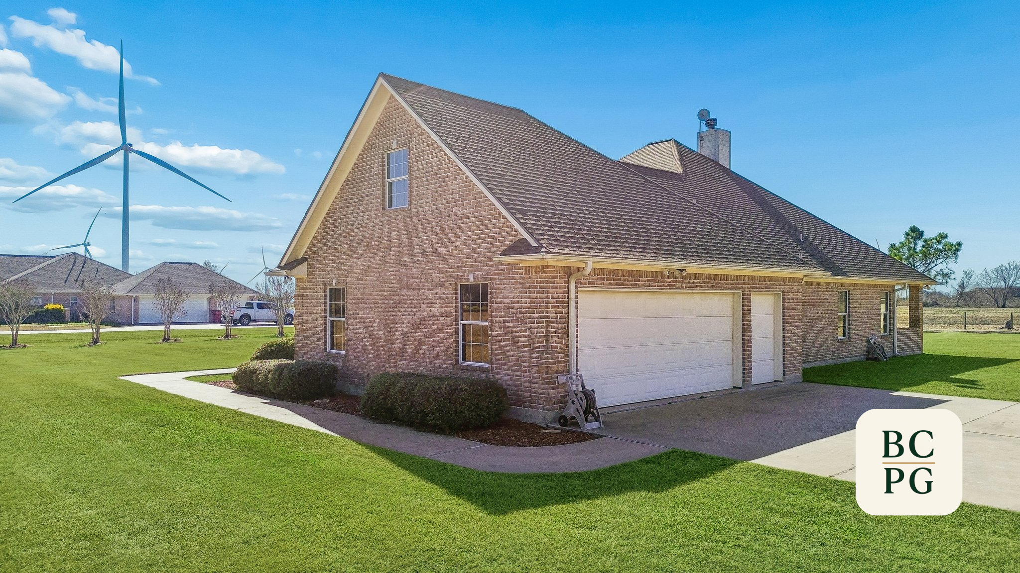 Front view of a brick house with a two-car garage, surrounded by a well-maintained lawn, in a suburban neighborhood with wind turbines and other houses in the background under a partly cloudy sky.