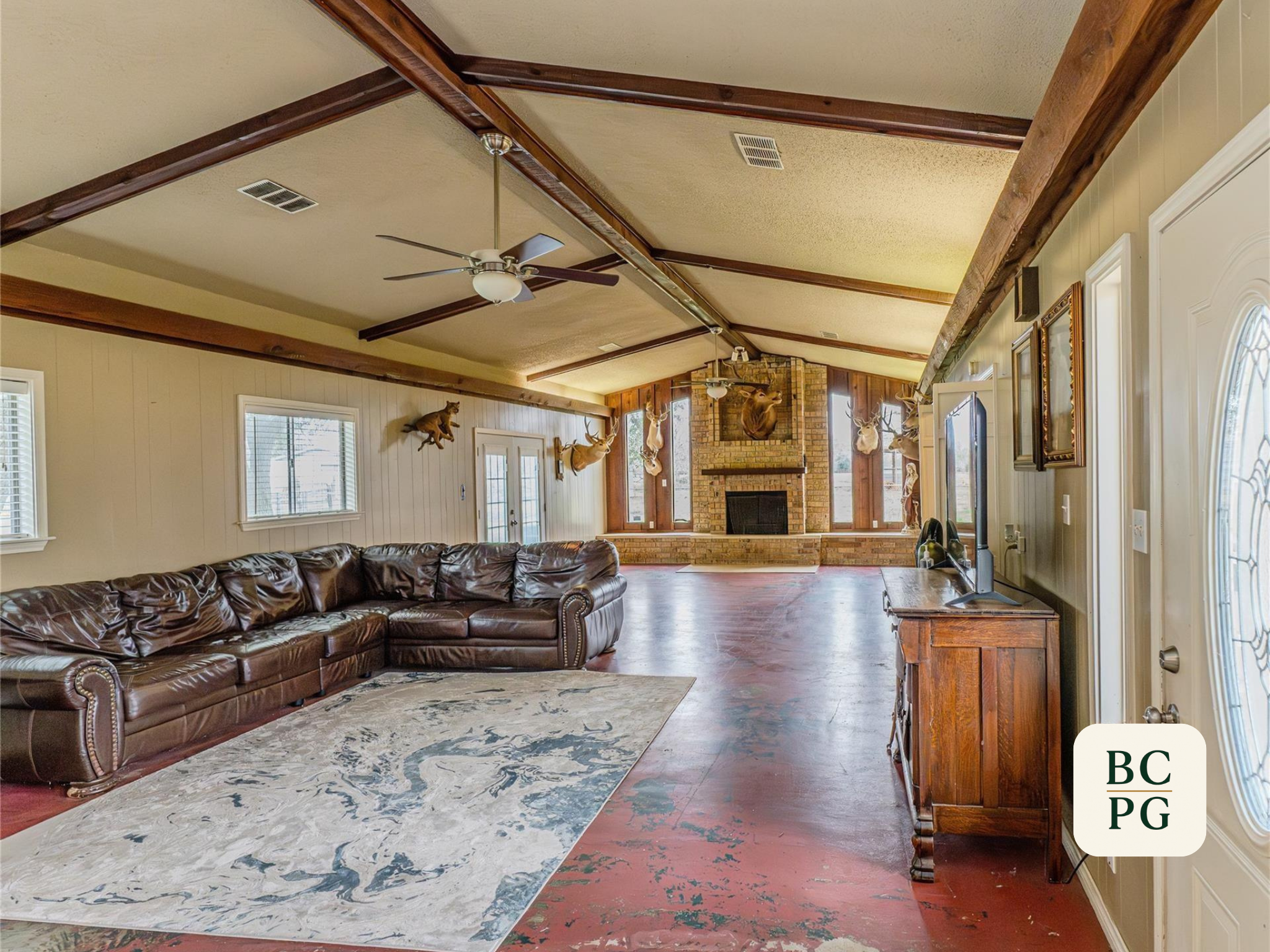 Living room with vaulted ceiling featuring exposed wooden beams, ceiling fans, mounted deer and animal heads, brick fireplace, windows, leather sectional sofa, and a decorative rug.