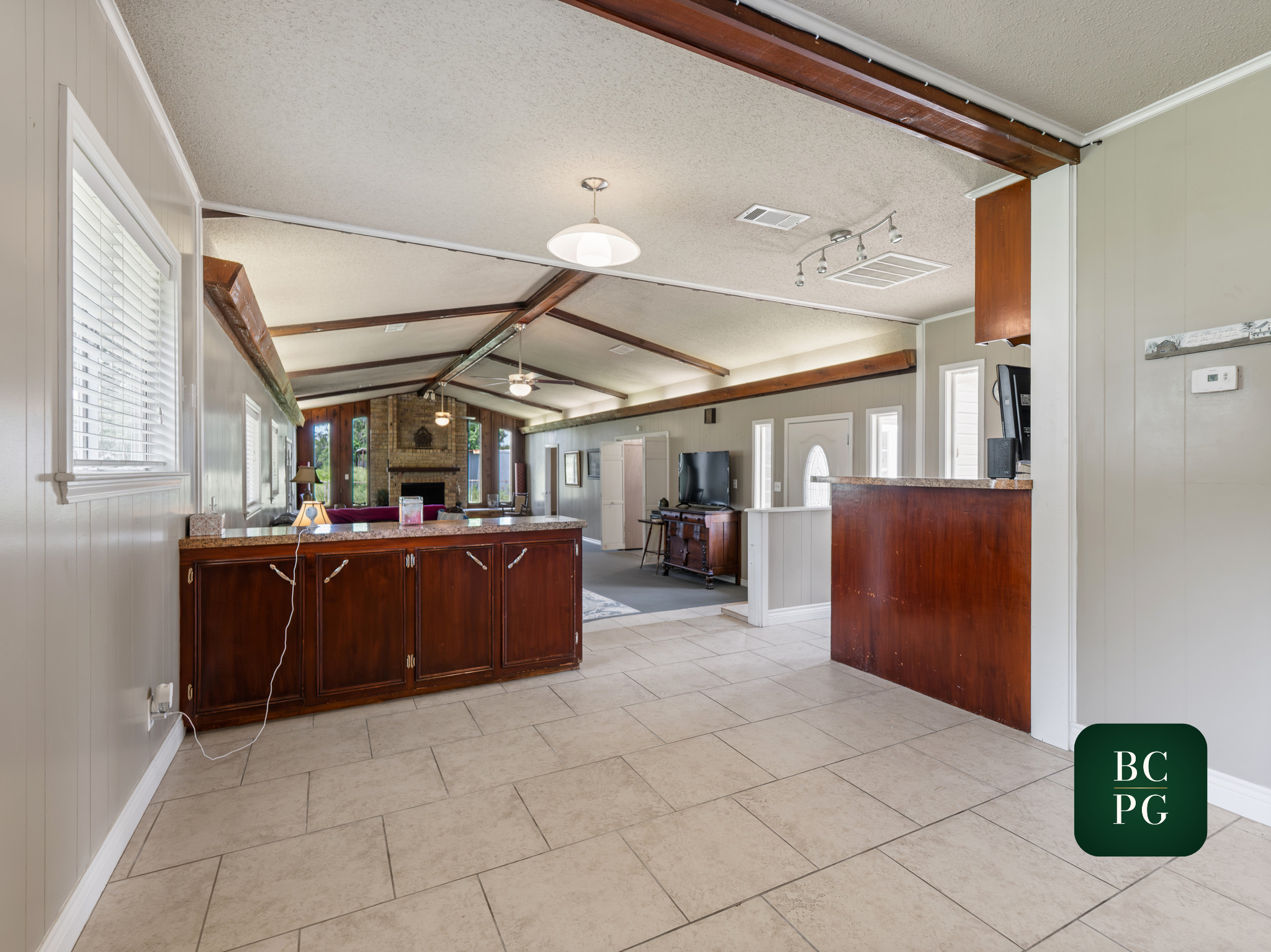 A spacious living room with a high, textured ceiling featuring exposed wooden beams and hanging lights. The room has several large windows allowing natural light, and includes a brick fireplace, a large flat-screen TV, and various furniture pieces.