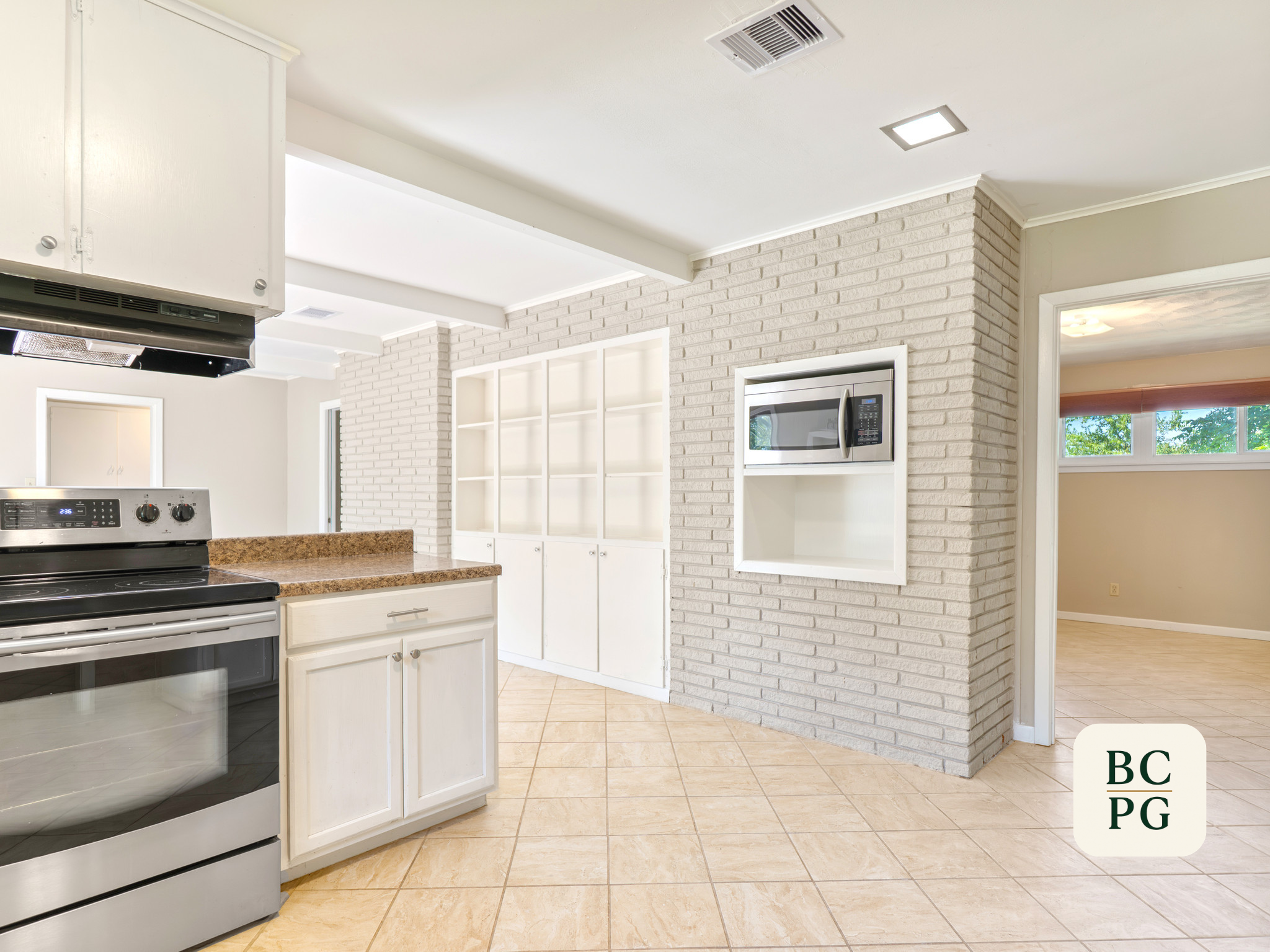 Modern kitchen with white cabinetry, brick wall, microwave, stove, and tiled floor, with a view into a room with a window and trees outside.