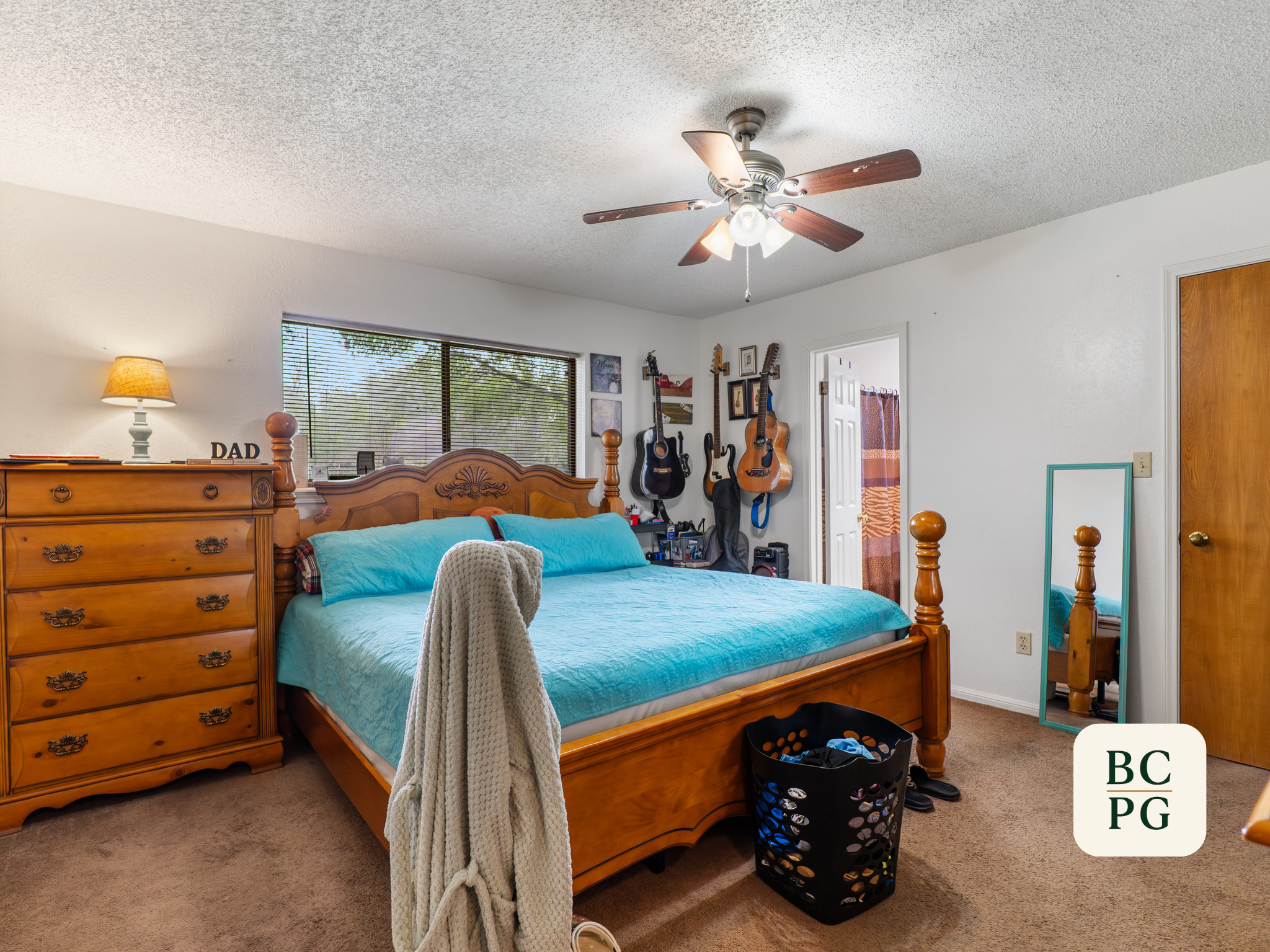 A bedroom with a wooden bed frame and teal bedding, guitar wall decor, a window with blinds, a dresser with a lamp, and a closet door, with a laundry basket on the carpeted floor.