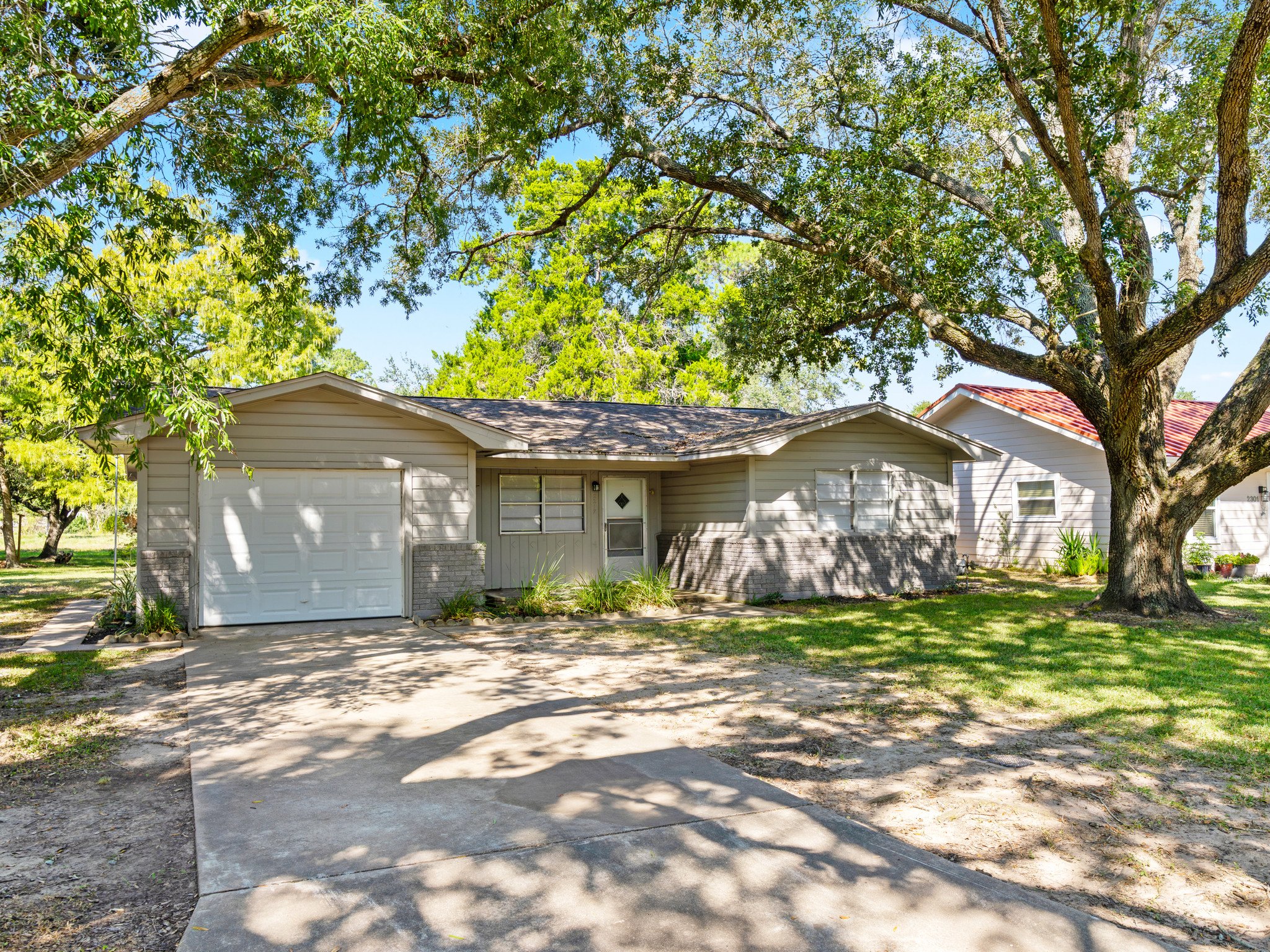 A single-story house with beige siding, a front lawn, and large trees providing shade. The house has a small porch and a white garage door. The driveway leads up to the garage, and there is a neighboring house visible to the right with a red roof.