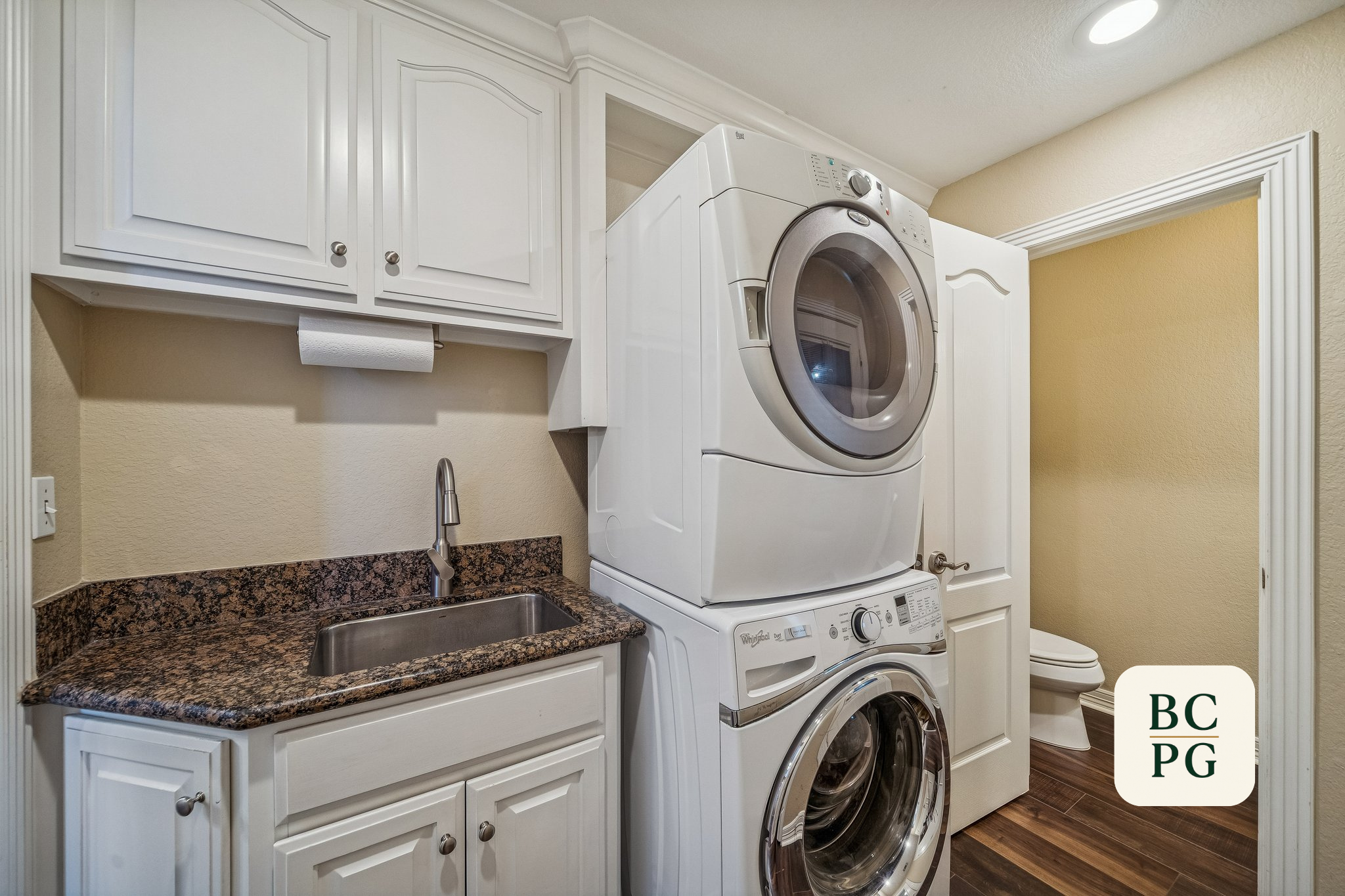 A laundry room with white cabinets, a granite countertop, a stainless steel sink, a Whirlpool washing machine stacked on top of a dryer, a paper towel holder, a small trash can, and a beige wall.