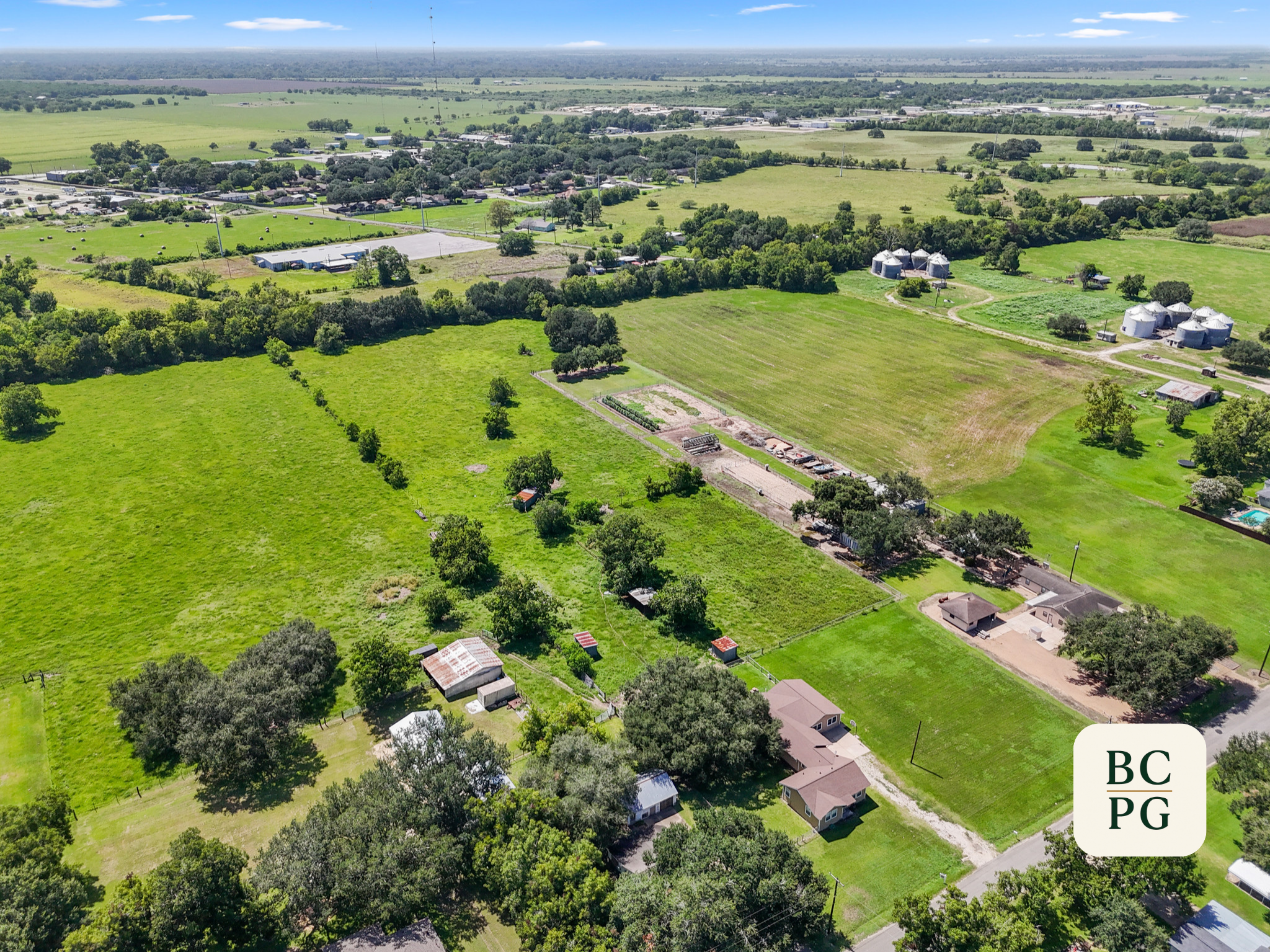 Aerial view of a rural landscape featuring green fields, trees, small houses, farm buildings, and silos under a partly cloudy sky.