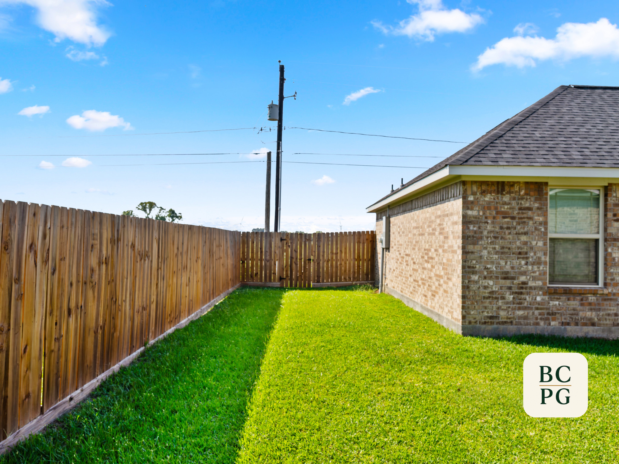 A backyard with a green lawn, wooden fence, and a brick house with a shingled roof against a bright blue sky with scattered clouds.