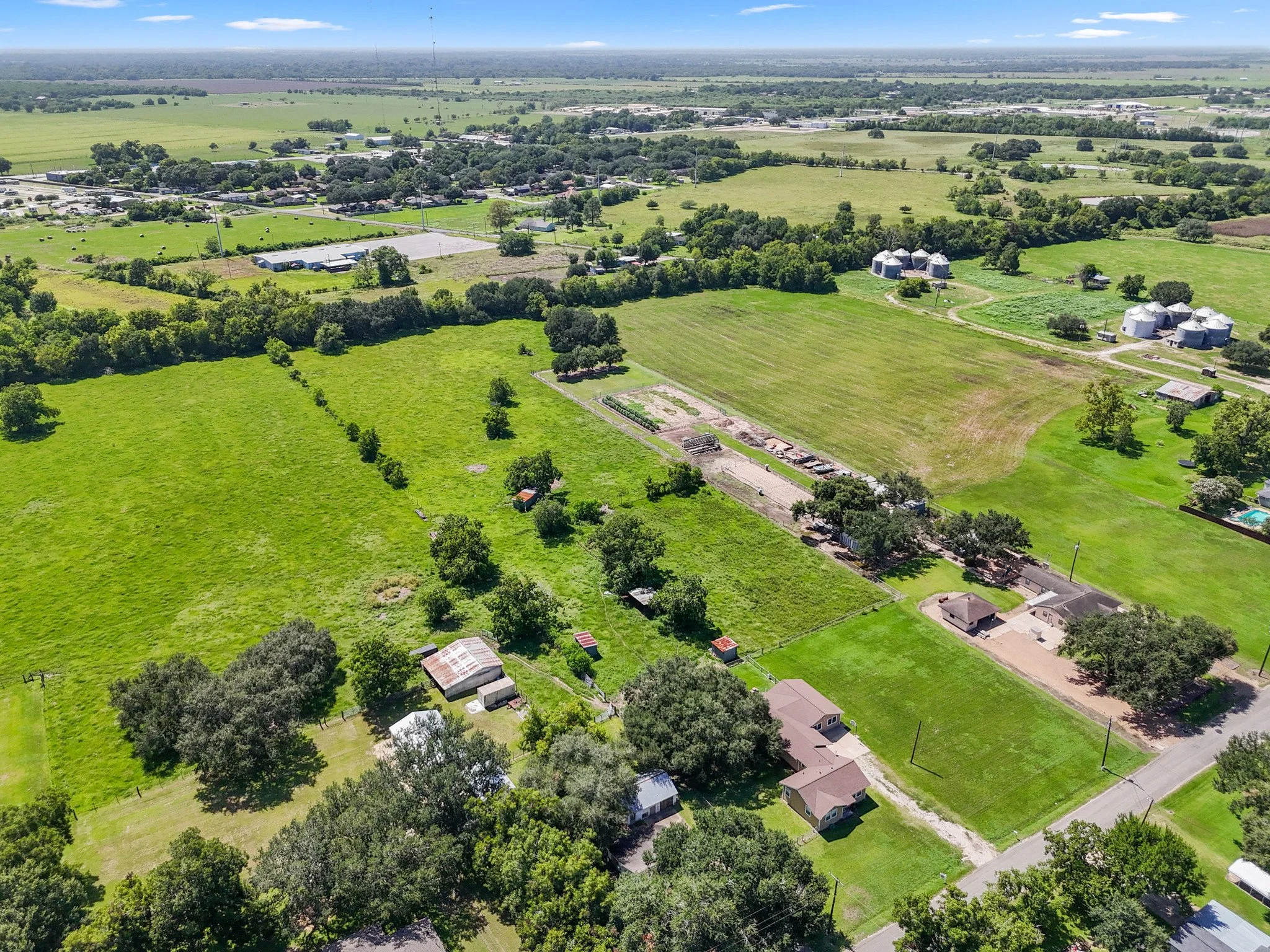 Aerial view of a rural area with green fields, trees, and scattered houses, some with sheds and barns, with a distant horizon under a partly cloudy sky.