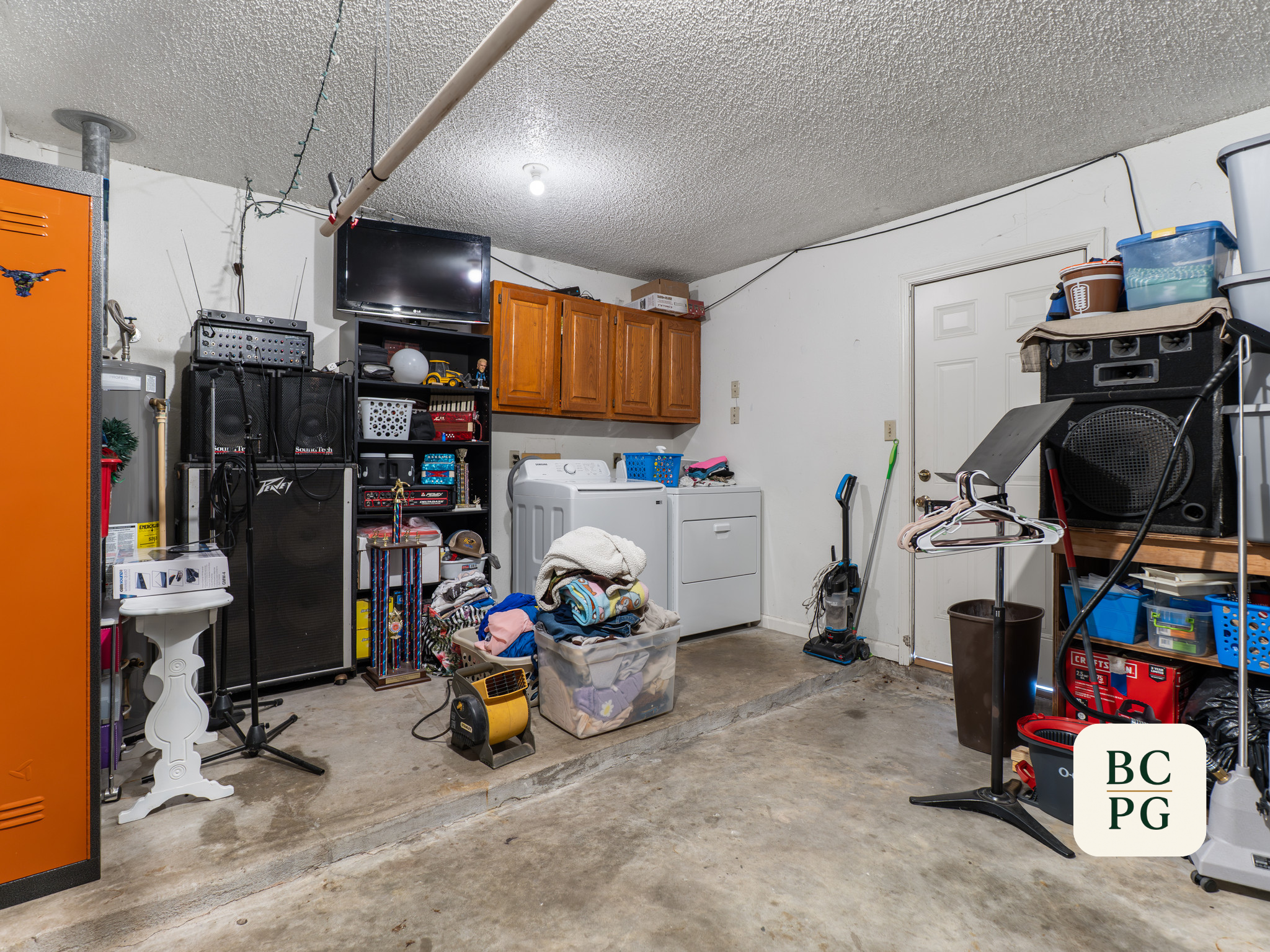 A cluttered garage with various household items, a washing machine, a dryer, shelves with containers, and a television mounted on the wall.