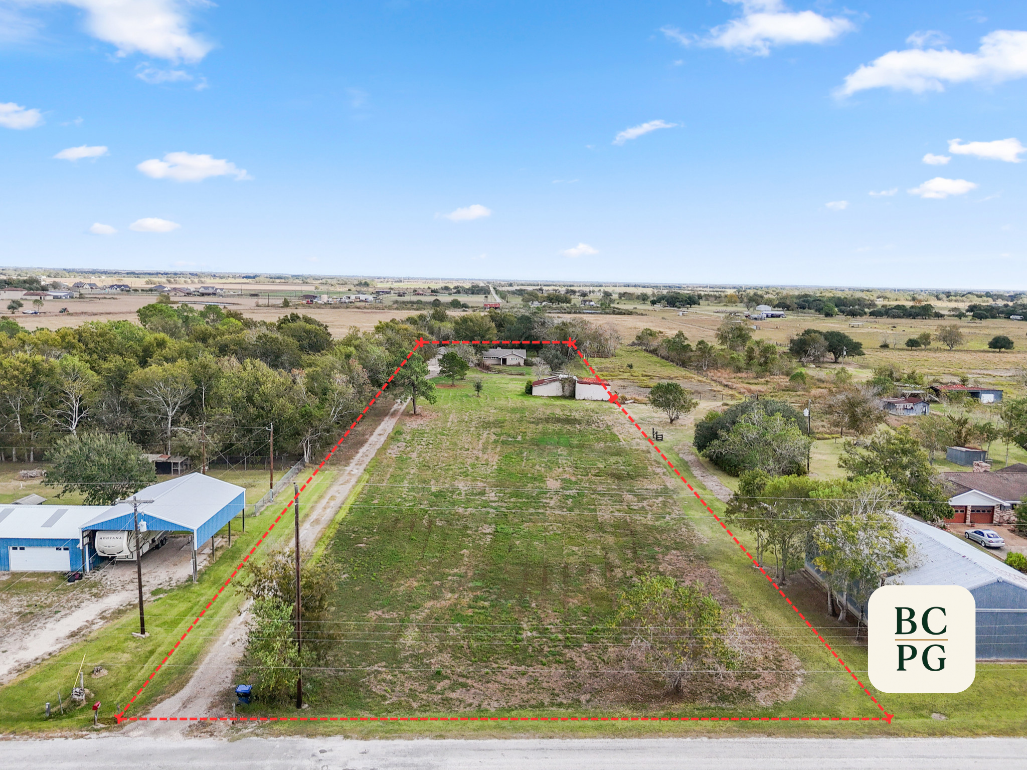 Aerial view of a rural property with a large open grassy area, trees, houses, and farm buildings under a blue sky with scattered clouds.