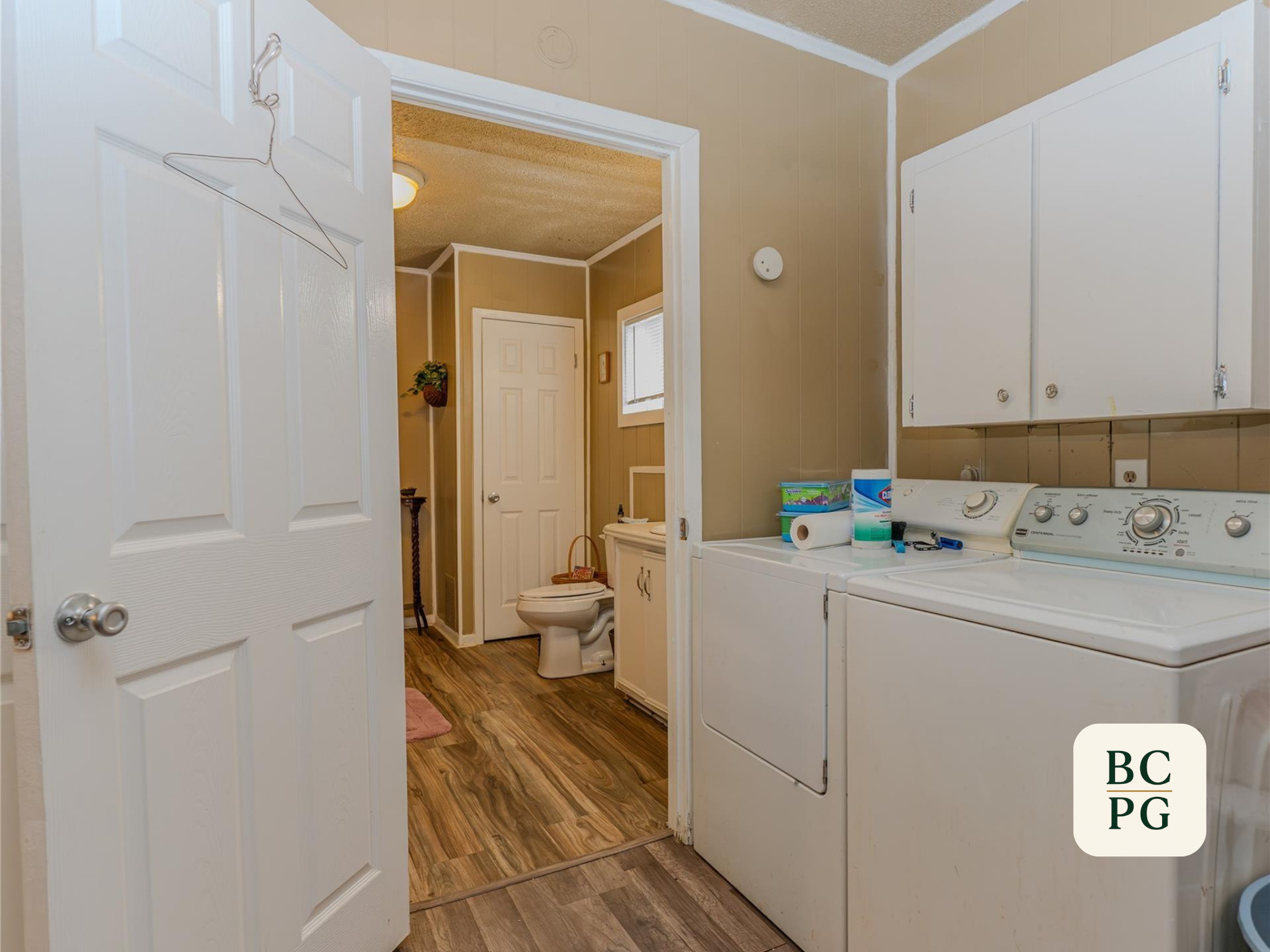 A view of a laundry room with white washer and dryer, beige walls, and wooden flooring. There are cabinets above the appliances and a small window in the background.