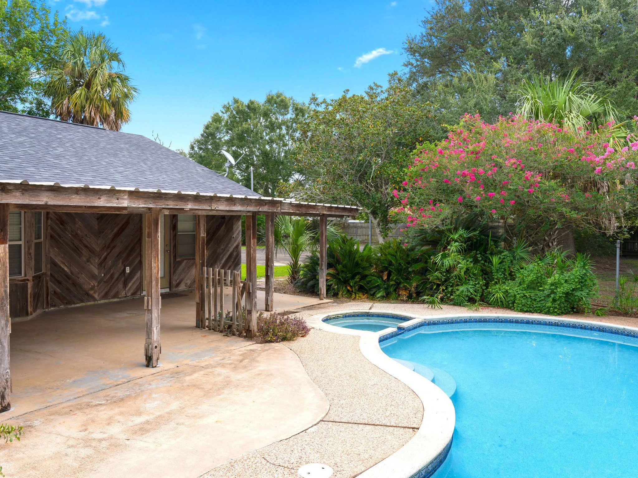 A backyard with a swimming pool, lush greenery, a covered patio area with wooden support beams, and flowering trees against a bright blue sky.