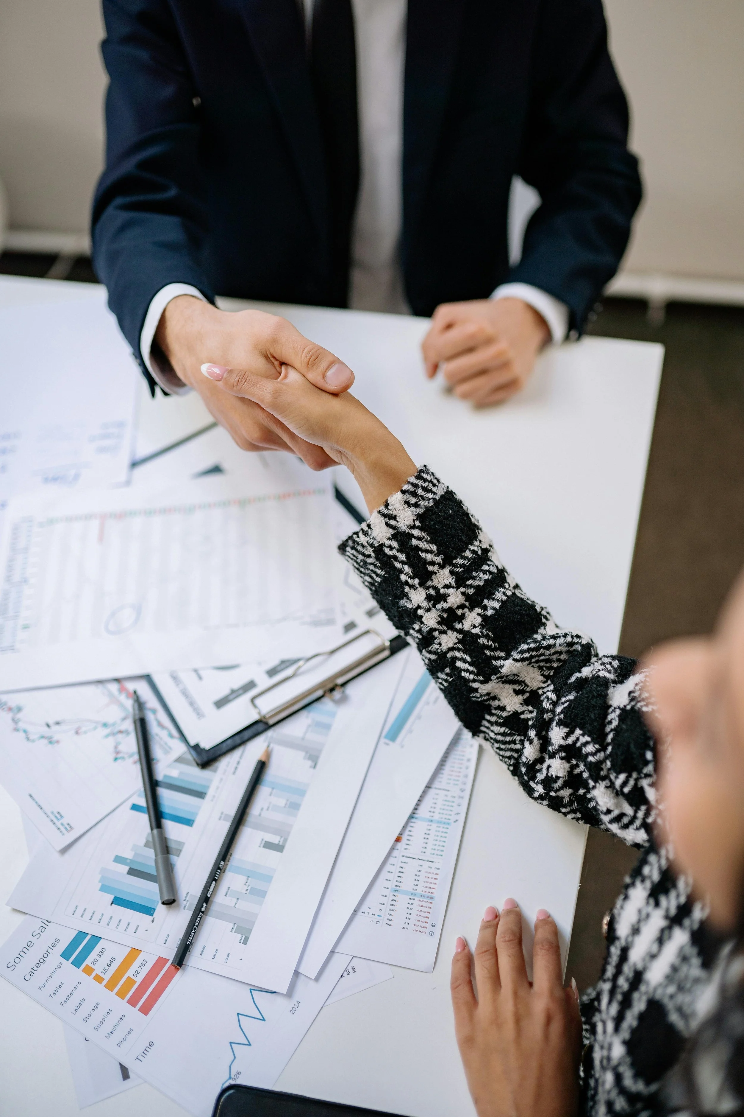 A person in a suit shaking hands with another person wearing a patterned sweater over a white shirt, in an office setting with documents and charts on the table.