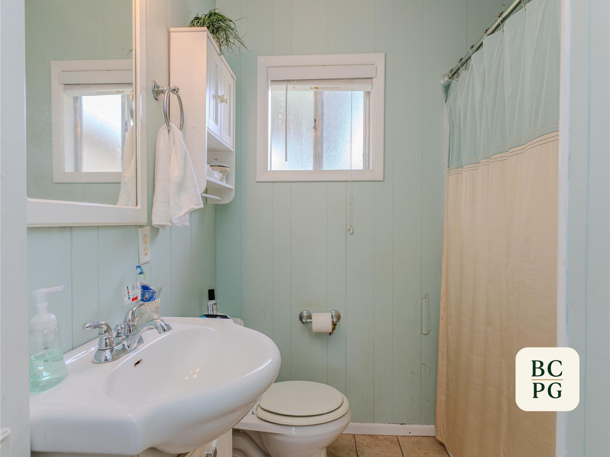 A small bathroom with light green walls, a white porcelain sink with soap and hand sanitizer, a mirror, a window with frosted glass, a toilet, a white cabinet, and a shower with a curtain.