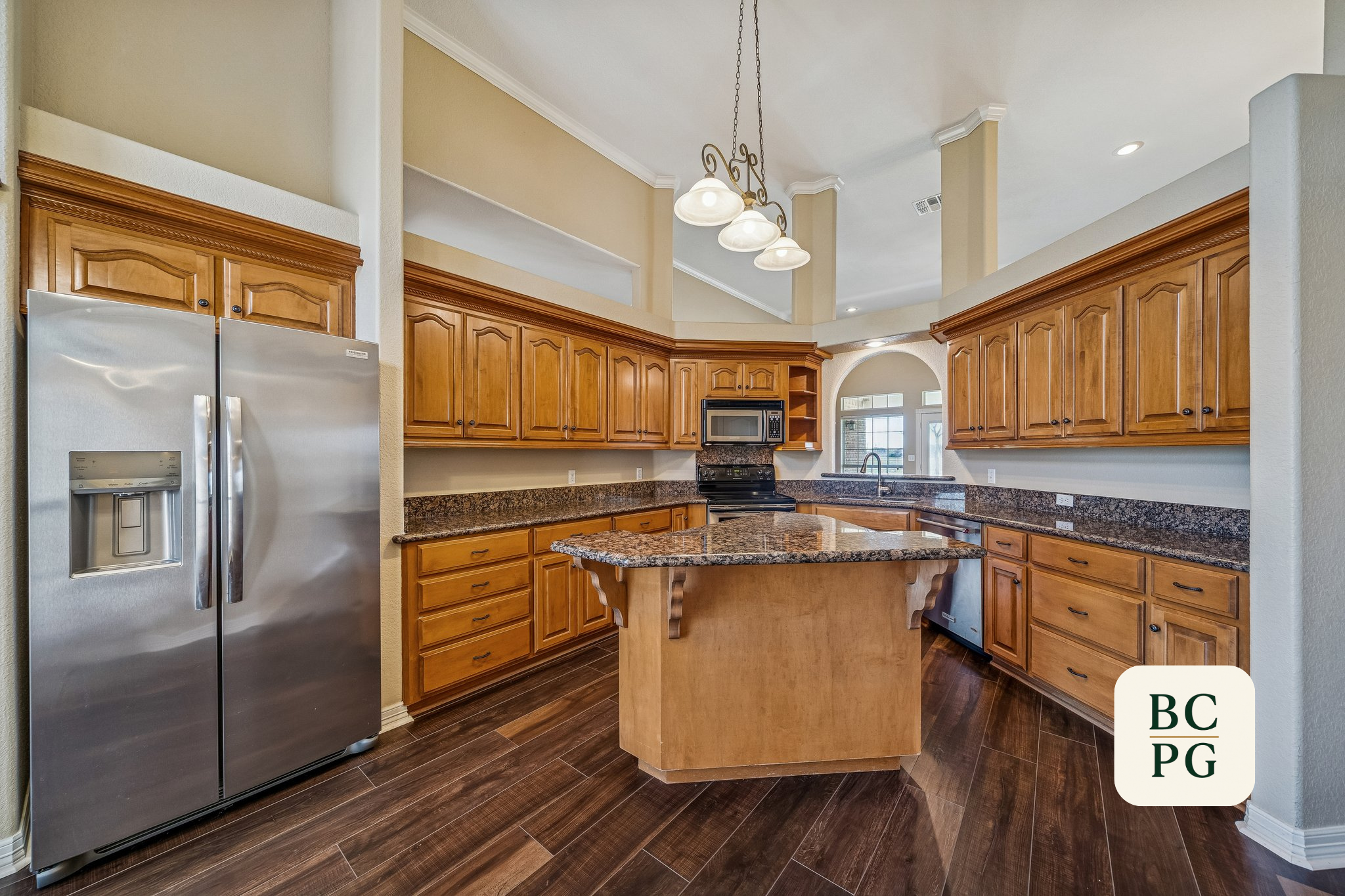 Kitchen with wooden cabinets, a granite island, stainless steel refrigerator, and a microwave, with a high ceiling and pendant lighting.
