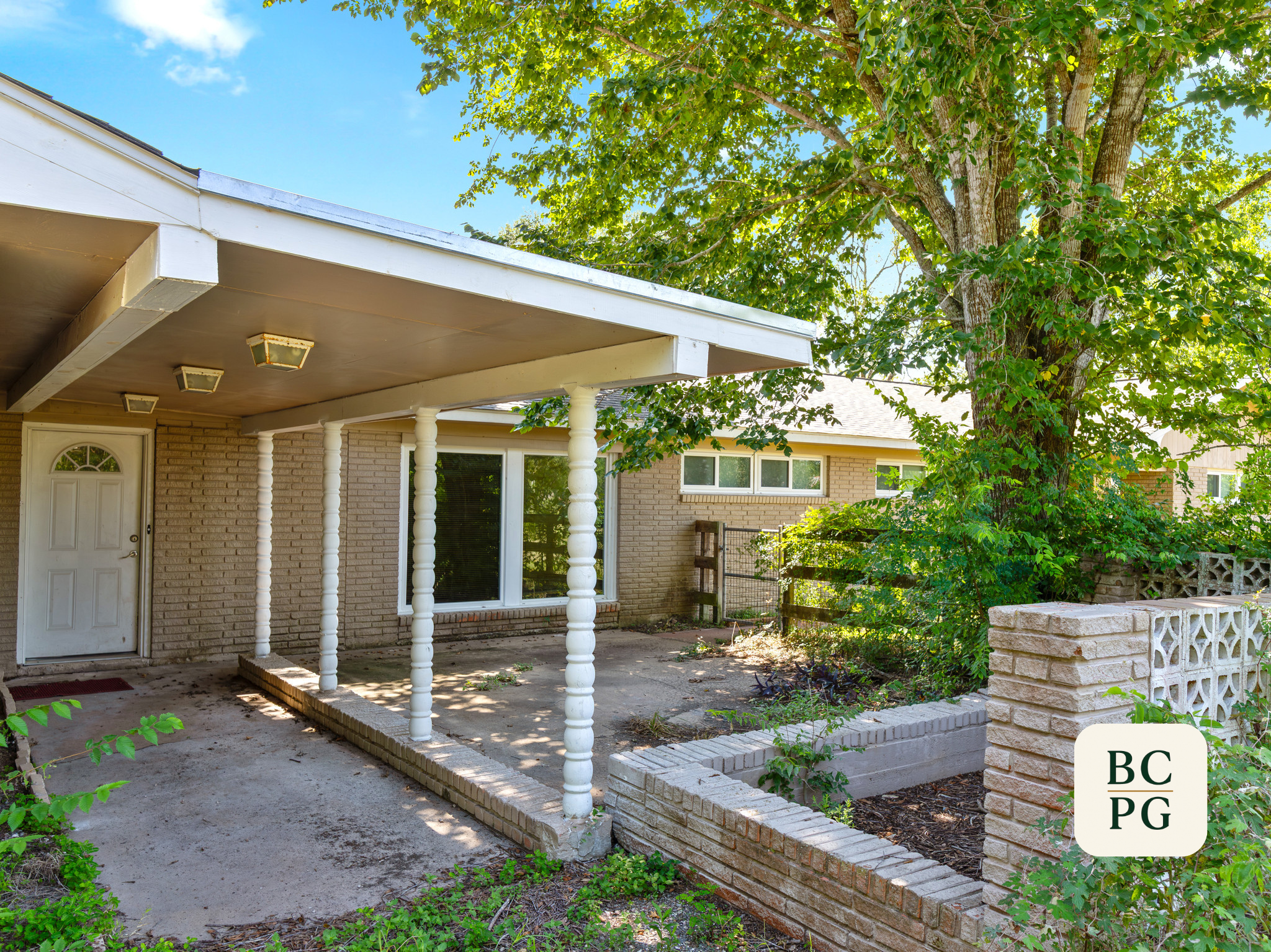 Front porch area of a house with white columns supporting a roof extension, a brick wall, a door, and windows, with a large leafy tree and a brick planter in the garden.