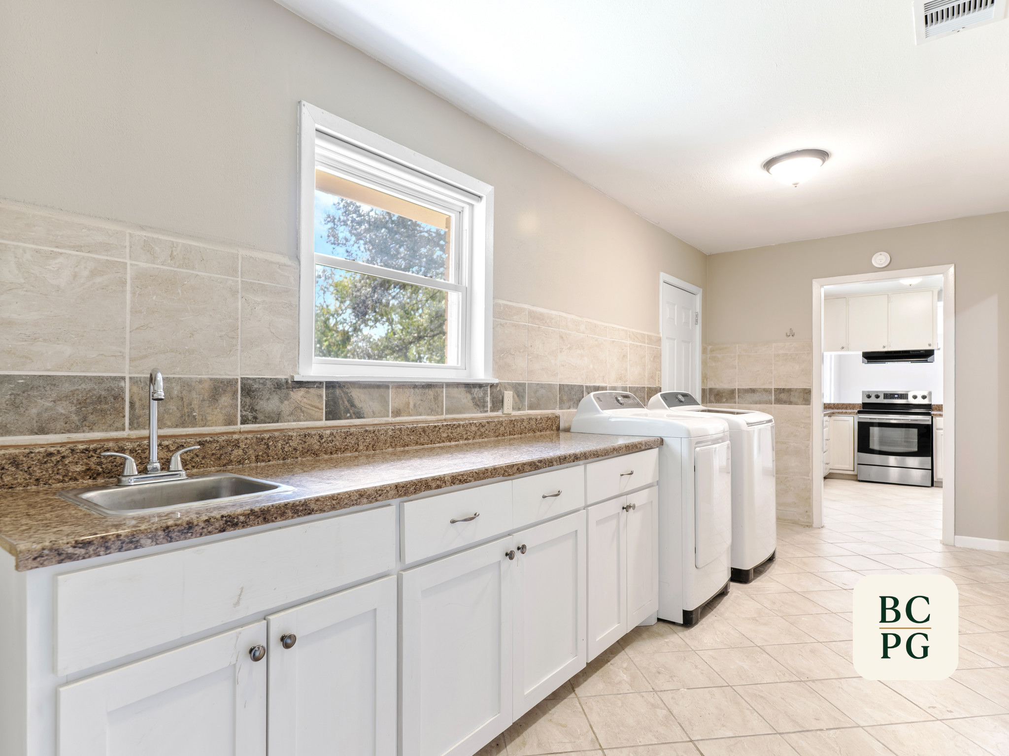 A laundry room with a granite countertop, white cabinets, a sink, a window showing trees outside, and two top-loading washing machines. There's also a stove and oven visible through a doorway.