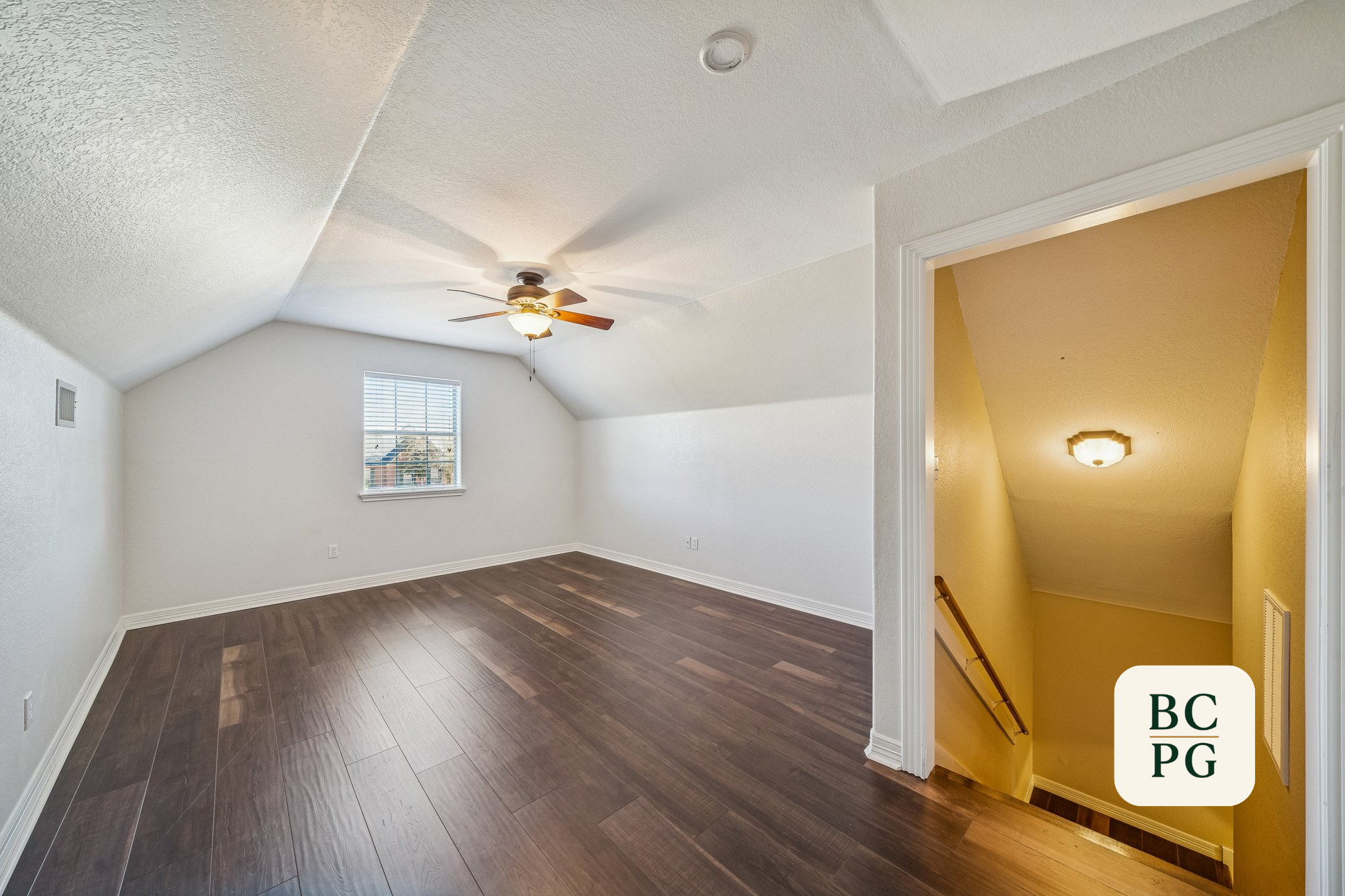 Empty room with white walls, hardwood flooring, a ceiling fan with a light, and a window with blinds. A hall with yellow walls and a light fixture is visible through a doorway.