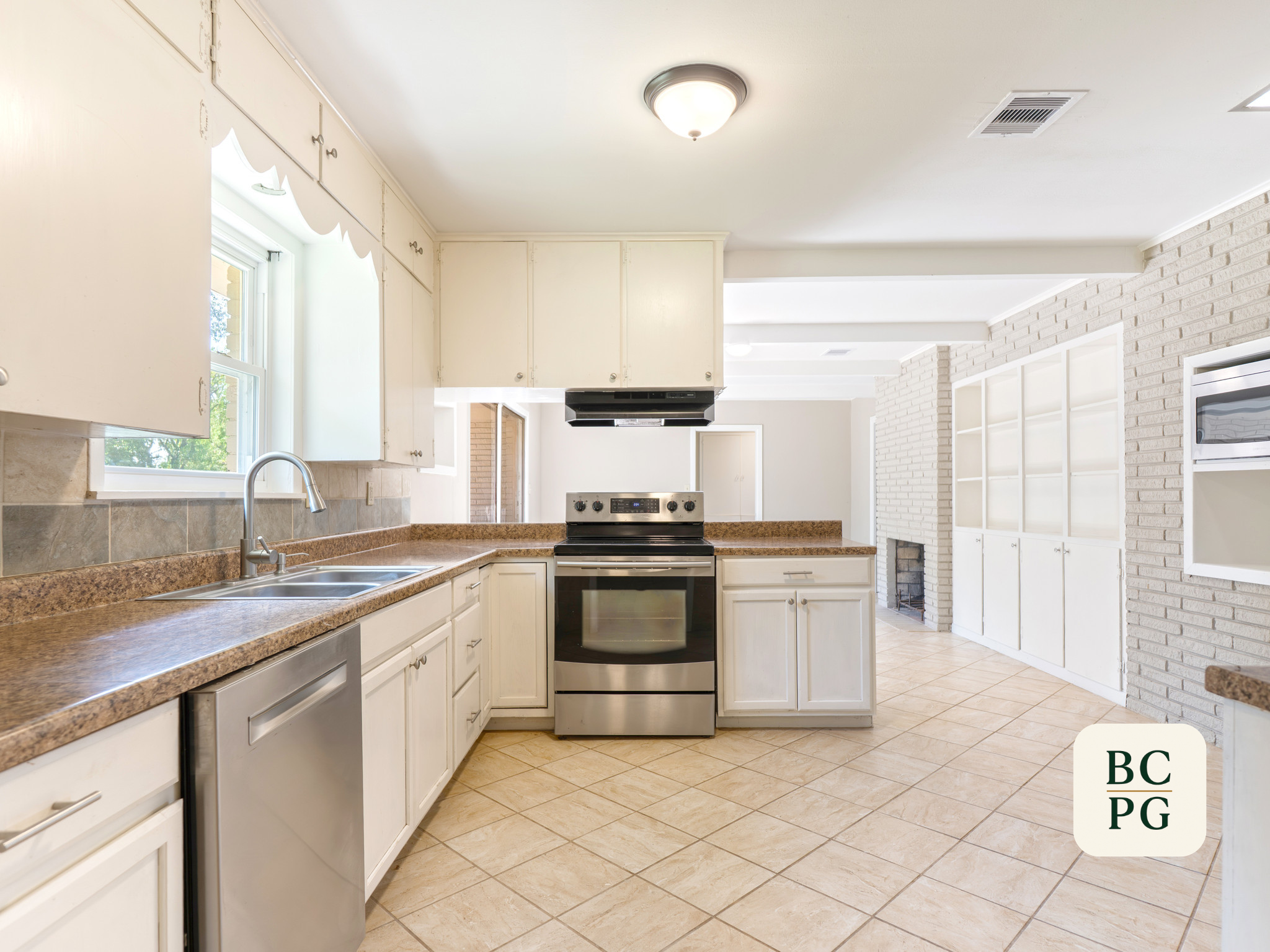 A kitchen with white cabinets, a brown countertop, a stainless steel oven and stove, a stainless steel sink, and a window letting in natural light. The floor has beige tiles, and there is a brick accent wall on the right.